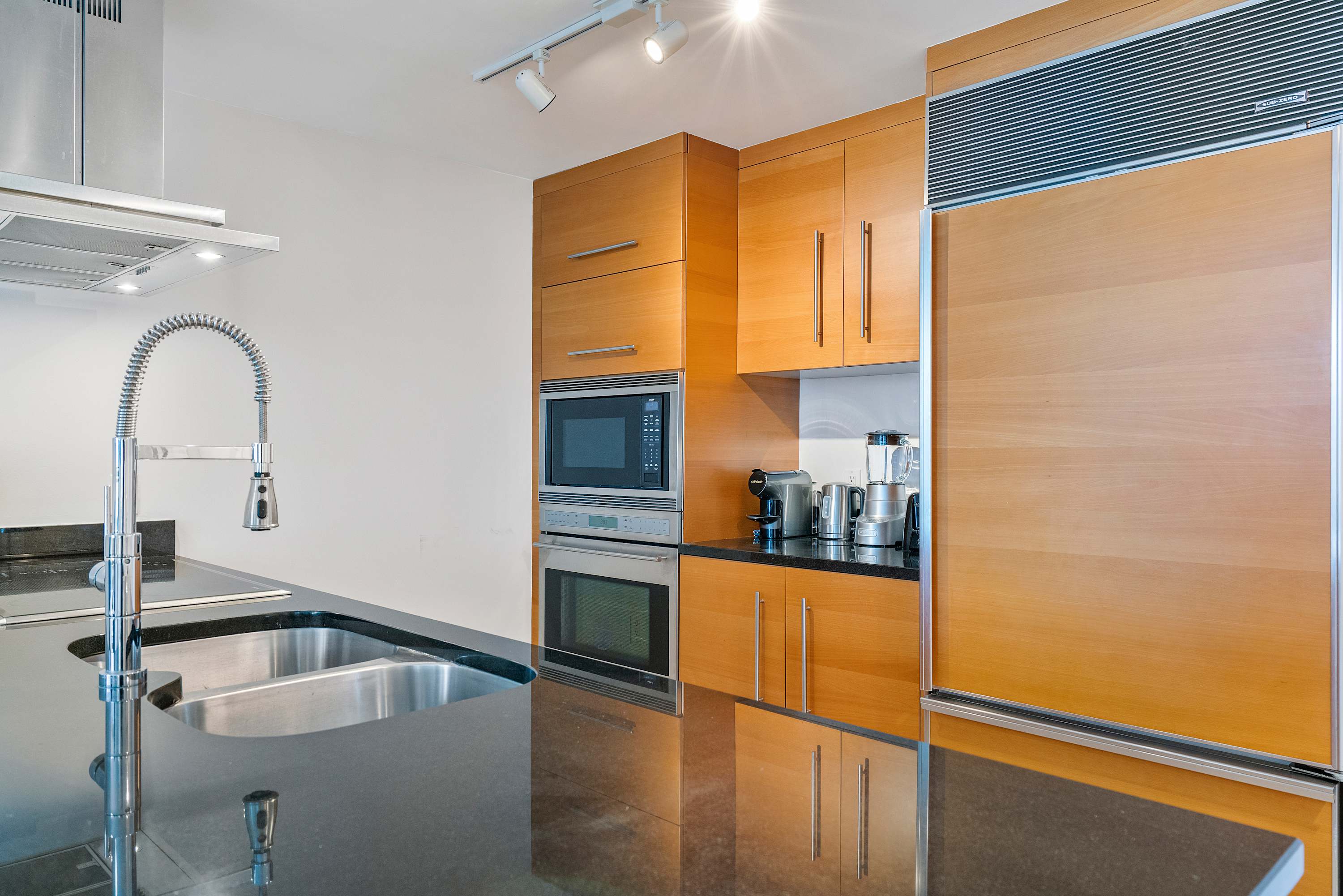 Modern kitchen featuring dark granite countertops, light wood cabinetry, stainless steel appliances including a built-in microwave and oven, and a large sink with a gooseneck faucet.