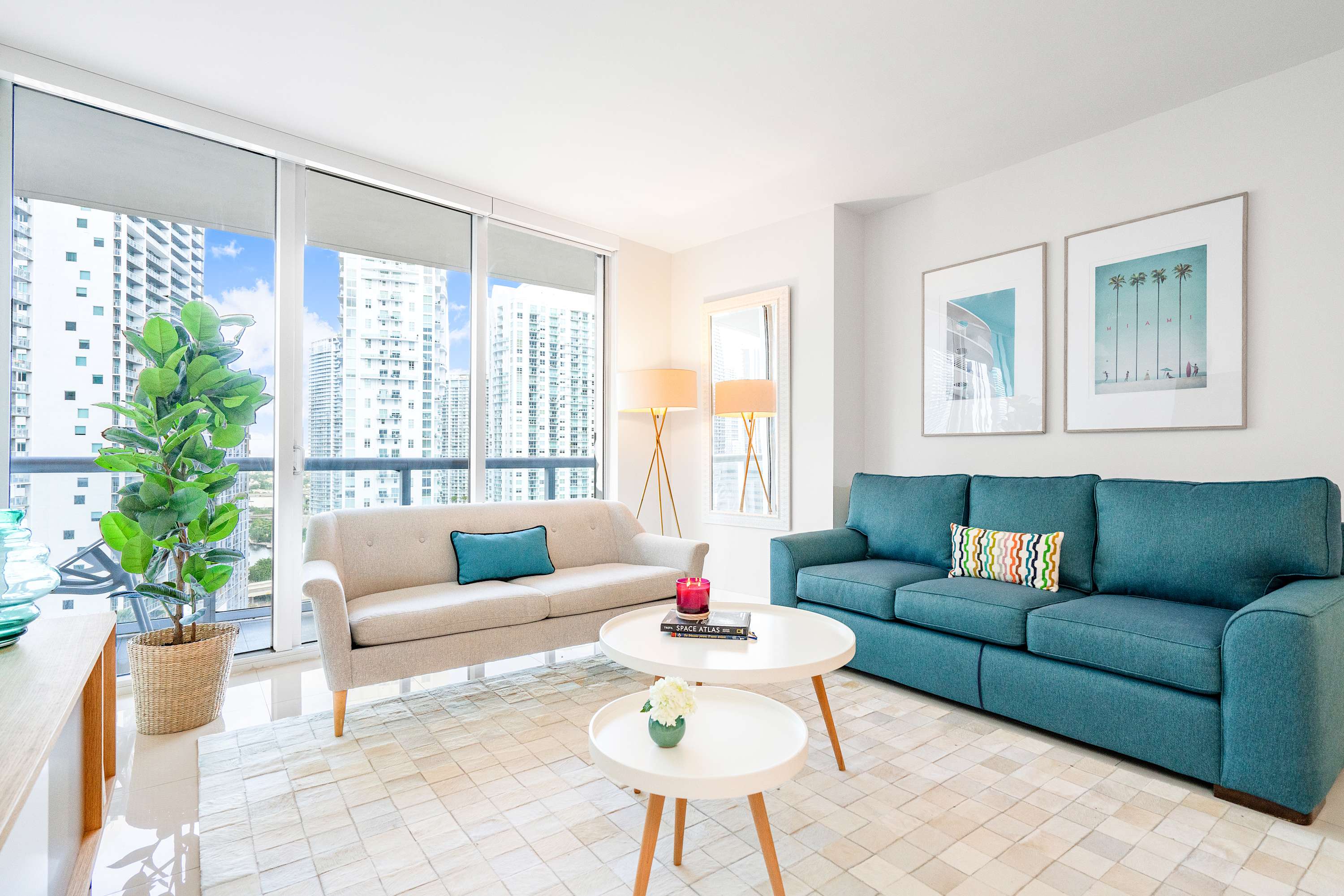 Spacious living room featuring a teal sofa and a beige sofa, two round white coffee tables, and large windows with city views. The room has light-colored flooring and modern decor.