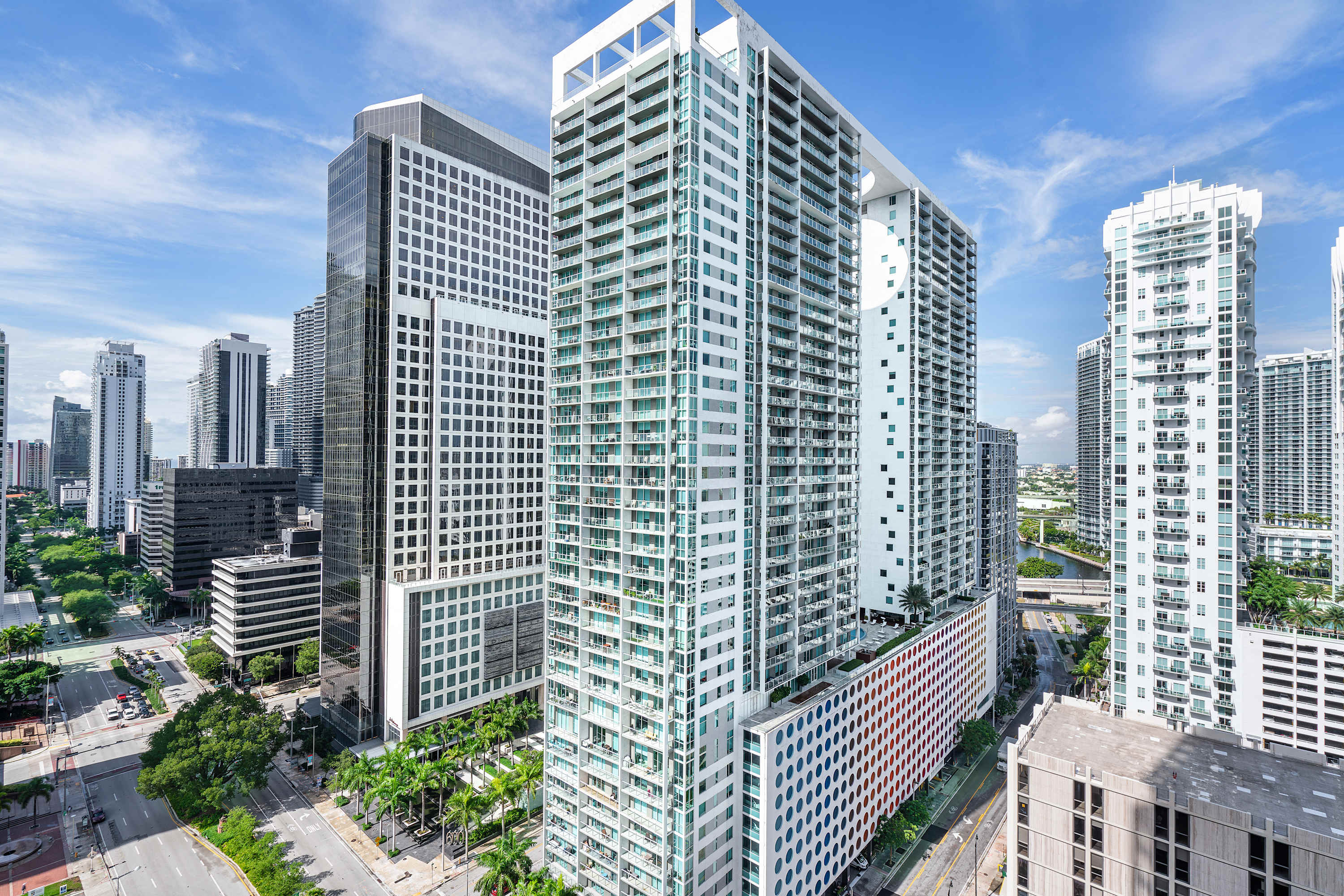 An expansive aerial view of a vibrant urban skyline dominated by numerous modern high-rise buildings with glass facades and balconies, surrounded by green spaces and city streets. A body of water is visible in the background.