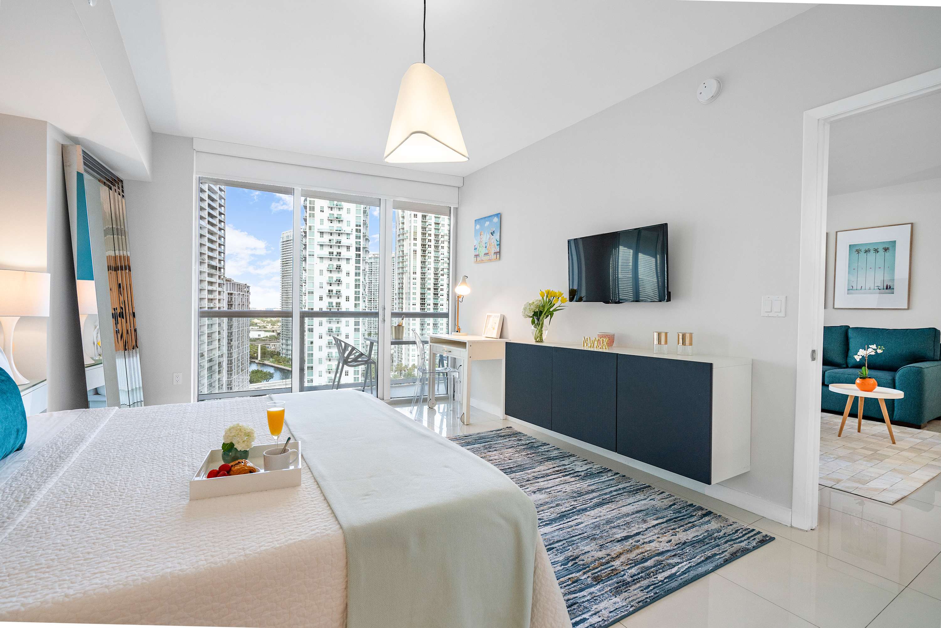 Bright bedroom with a white bed, a wall-mounted TV above a floating console, a small desk, and floor-to-ceiling windows offering panoramic city views. A blue rug adds a pop of color.
