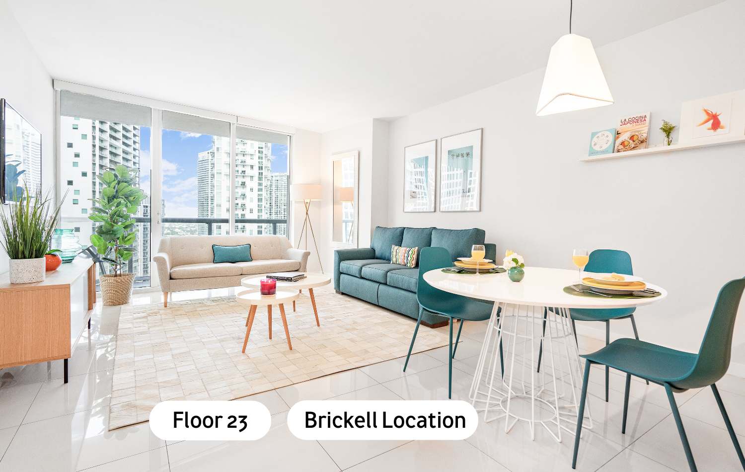 Bright, modern open-concept living and dining area with white tiled floors, a beige sofa, a teal sofa, two round coffee tables, and a white dining table with teal chairs, all bathed in natural light from large windows overlooking city buildings.