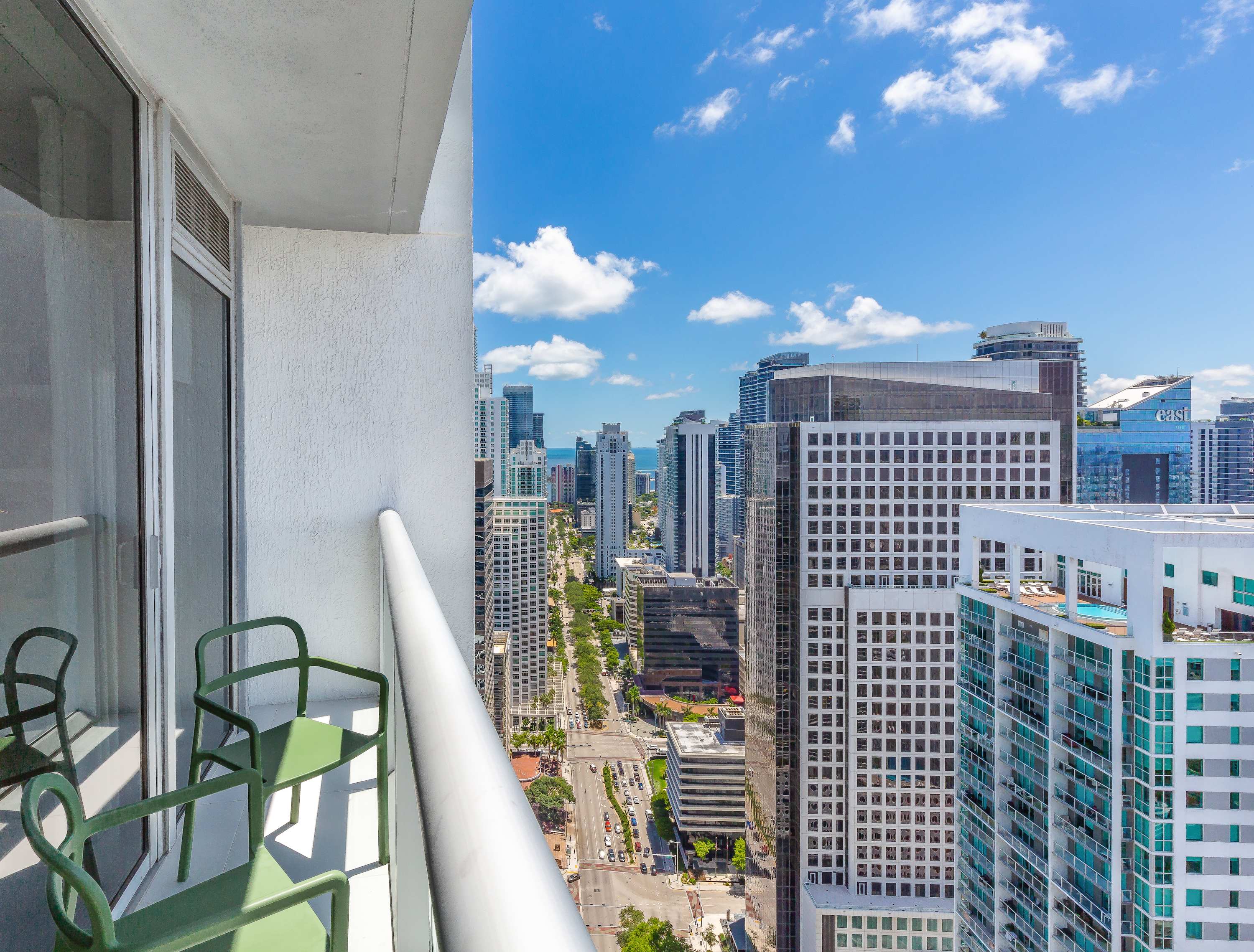 A high-rise balcony offers a panoramic city view with numerous skyscrapers, a street lined with cars, and the ocean visible in the distance under a bright blue sky with scattered clouds. Two green chairs and a small table are visible on the white balcony.