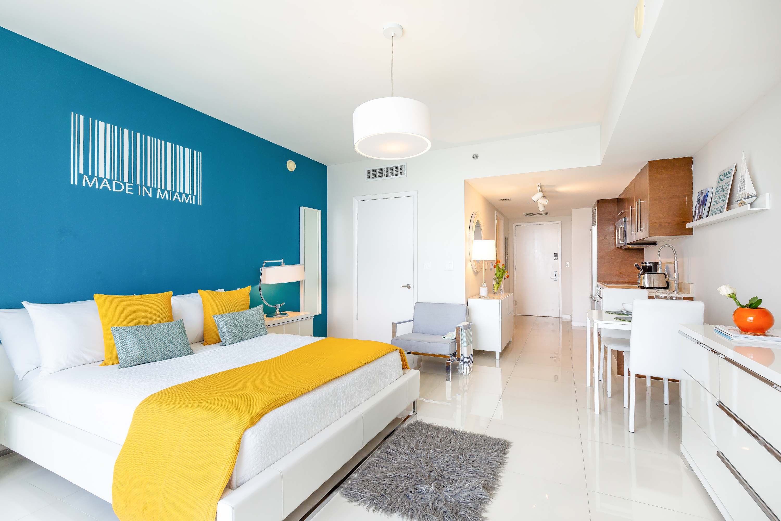 A modern studio apartment featuring a white bed with yellow and teal pillows against a striking blue accent wall with "MADE IN MIAMI" barcode art, an open living space, and a glimpse of the kitchen area.