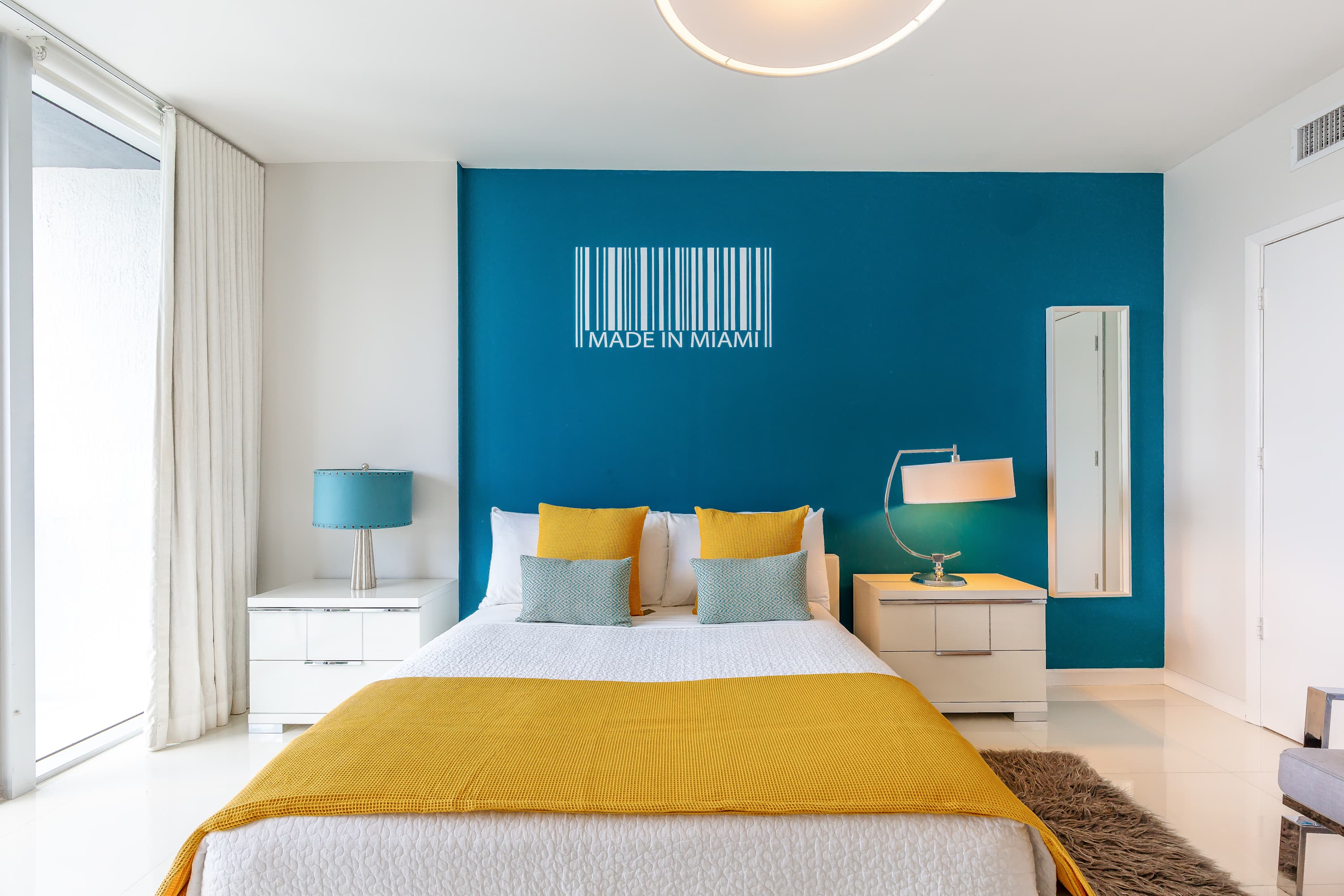 A bright bedroom area with a white bed, yellow and teal pillows, and a yellow throw blanket, set against a vibrant blue accent wall adorned with "MADE IN MIAMI" barcode art, with white nightstands and lamps.