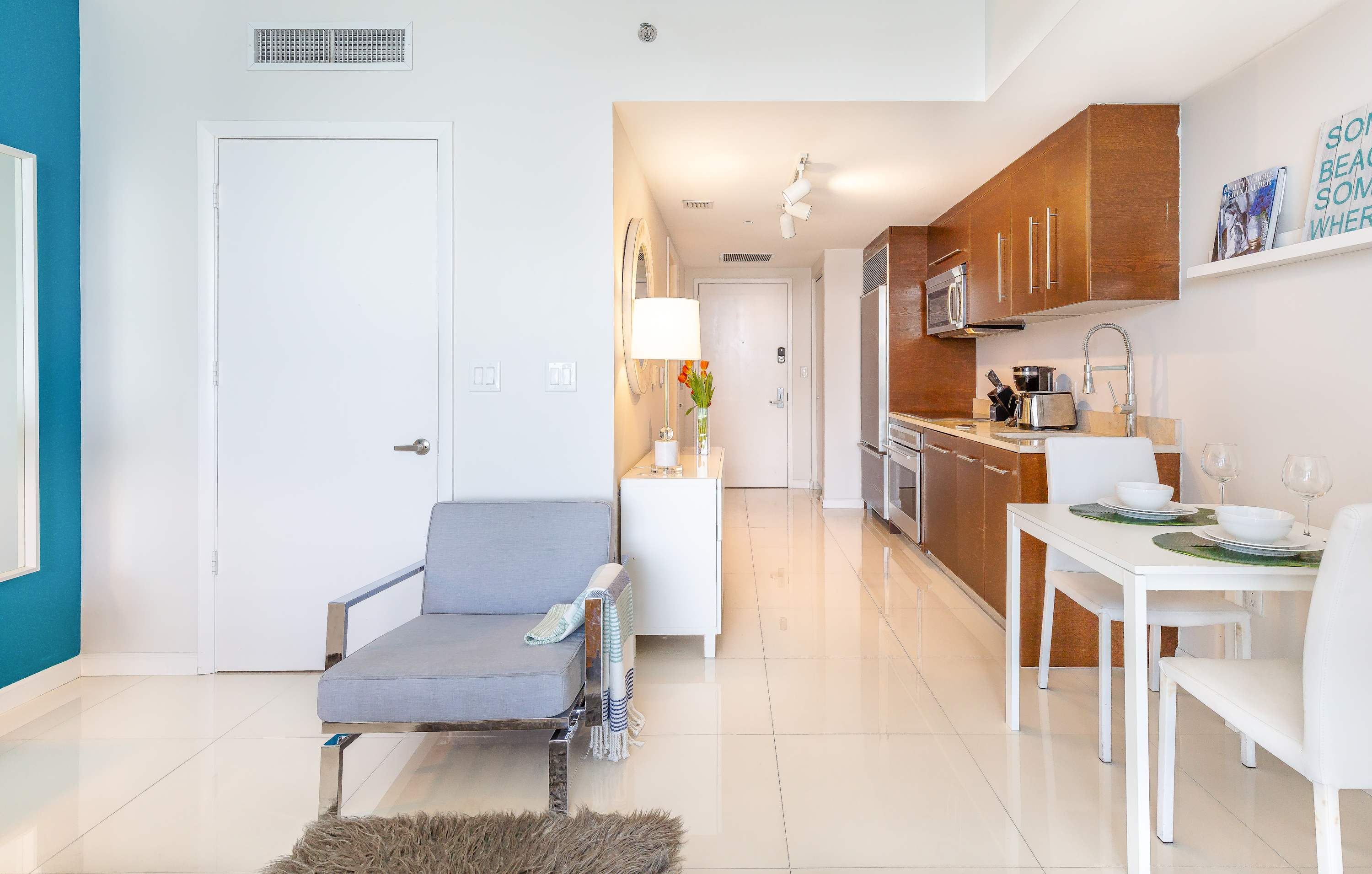 A sleek, open-plan living space with white tile flooring, featuring a grey chaise lounge, a white console table, and a compact kitchen with dark wood cabinetry and stainless steel appliances.