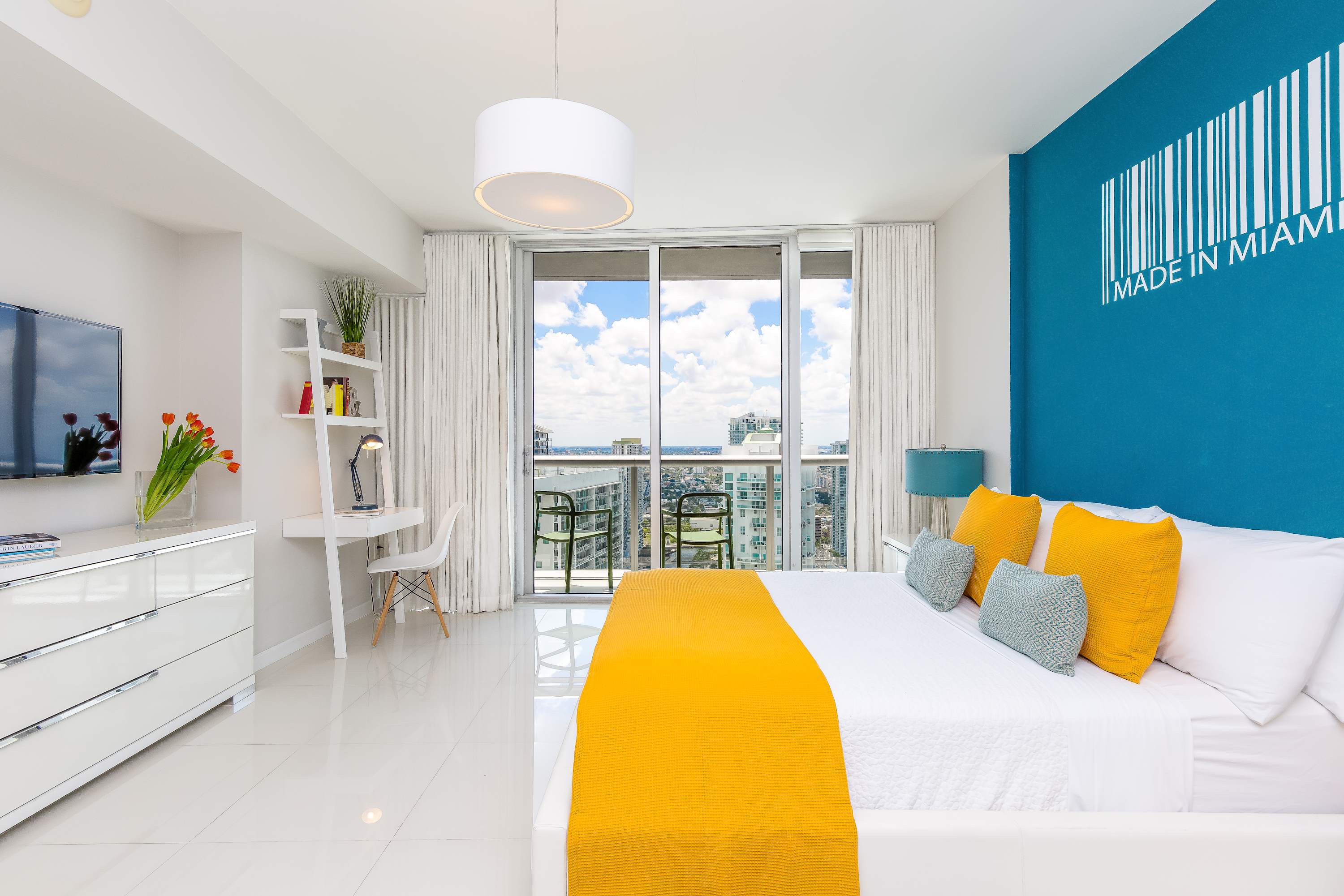 A bright and airy studio apartment with a white bed, a blue accent wall with "MADE IN MIAMI" art, a small desk area, and floor-to-ceiling windows leading to a balcony with city views.