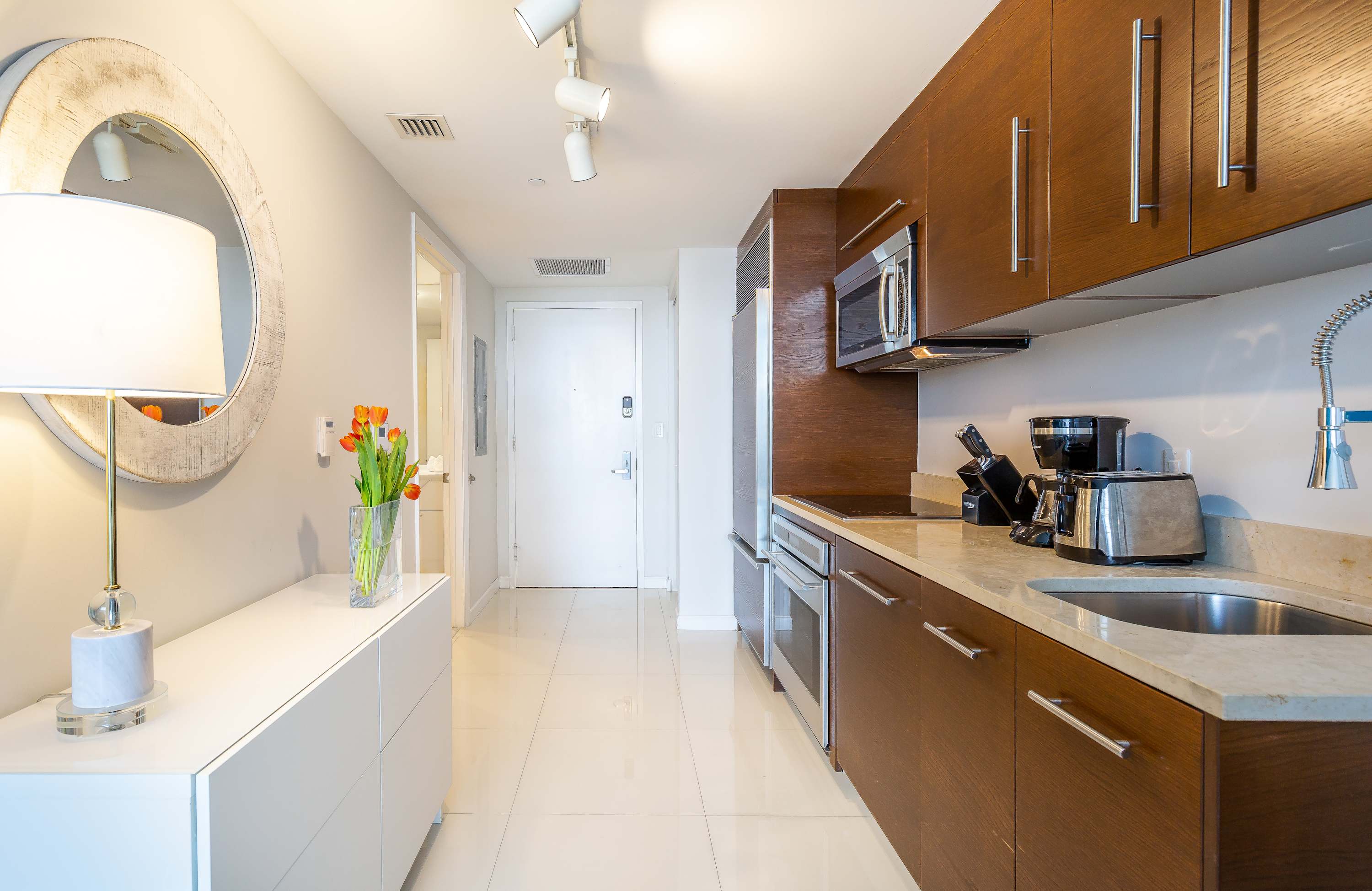 A compact, modern kitchen with dark wood cabinetry, light countertops, and stainless steel appliances, leading to a bright entryway with white tile flooring and a round mirror above a white console table.
