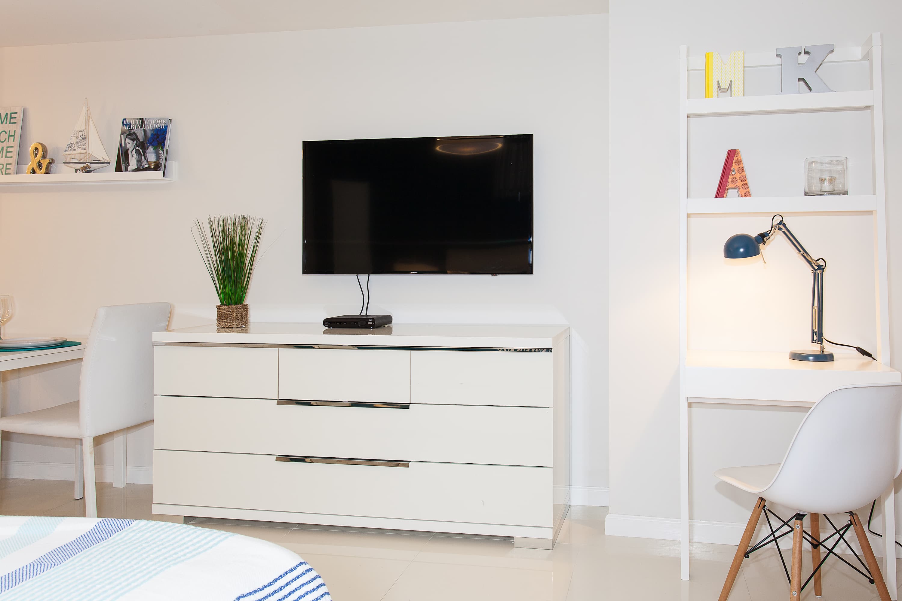 A contemporary living area featuring a wall-mounted flat-screen TV above a white dresser, a small white desk with a task lamp, and a white shelving unit, all on polished white tile flooring.
