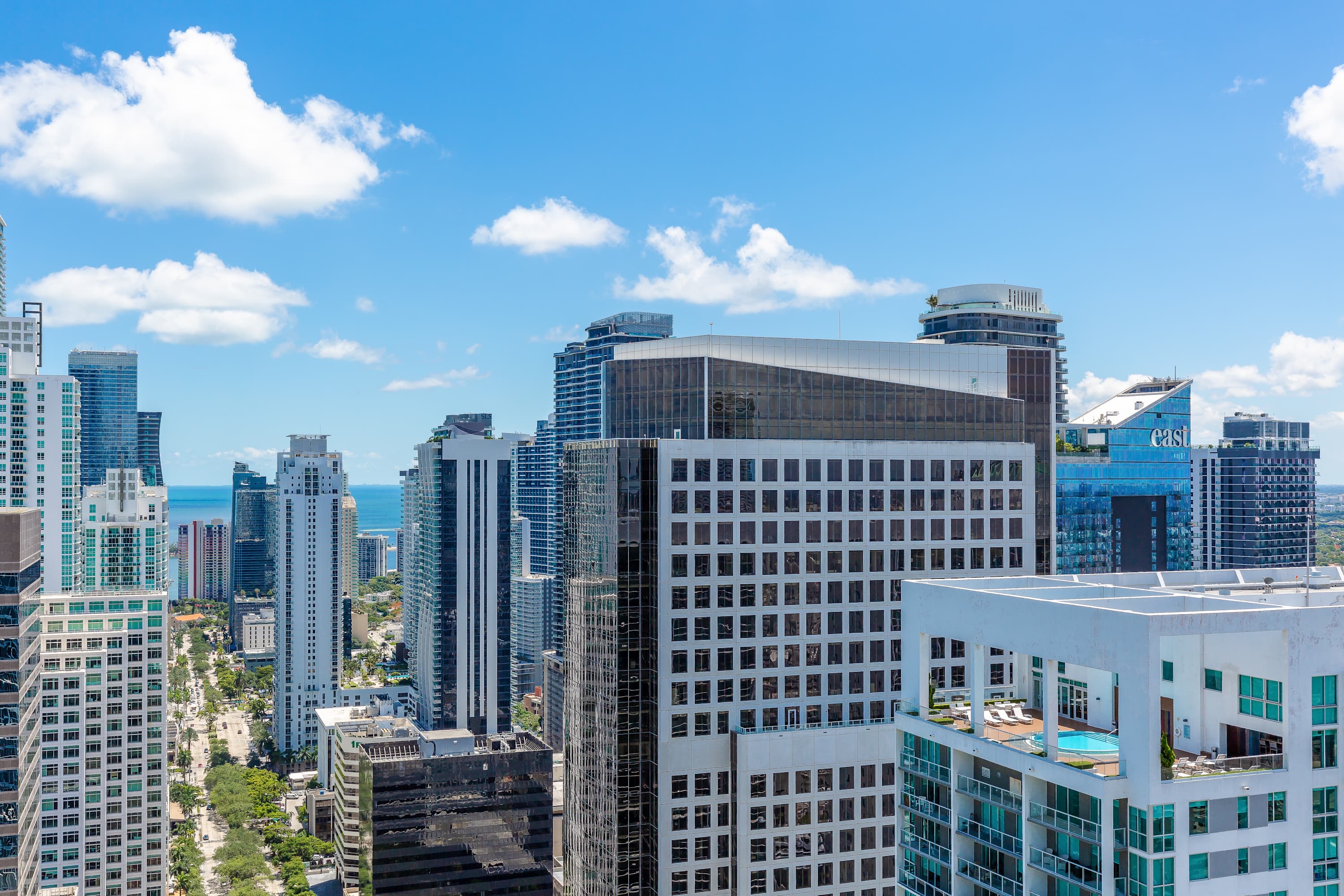 A panoramic city view from a high vantage point, featuring a dense cluster of modern high-rise buildings with varied architectural styles, extending towards the distant ocean under a partly cloudy blue sky.