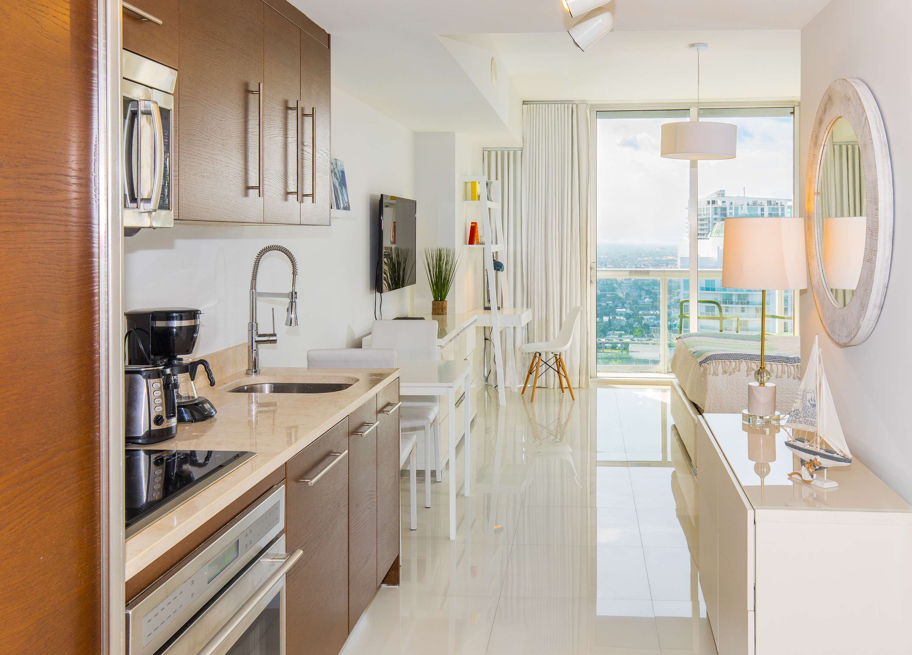 A modern kitchen with dark wood cabinets, light granite countertops, and stainless steel appliances, adjacent to a small white dining table, with large windows offering city views.