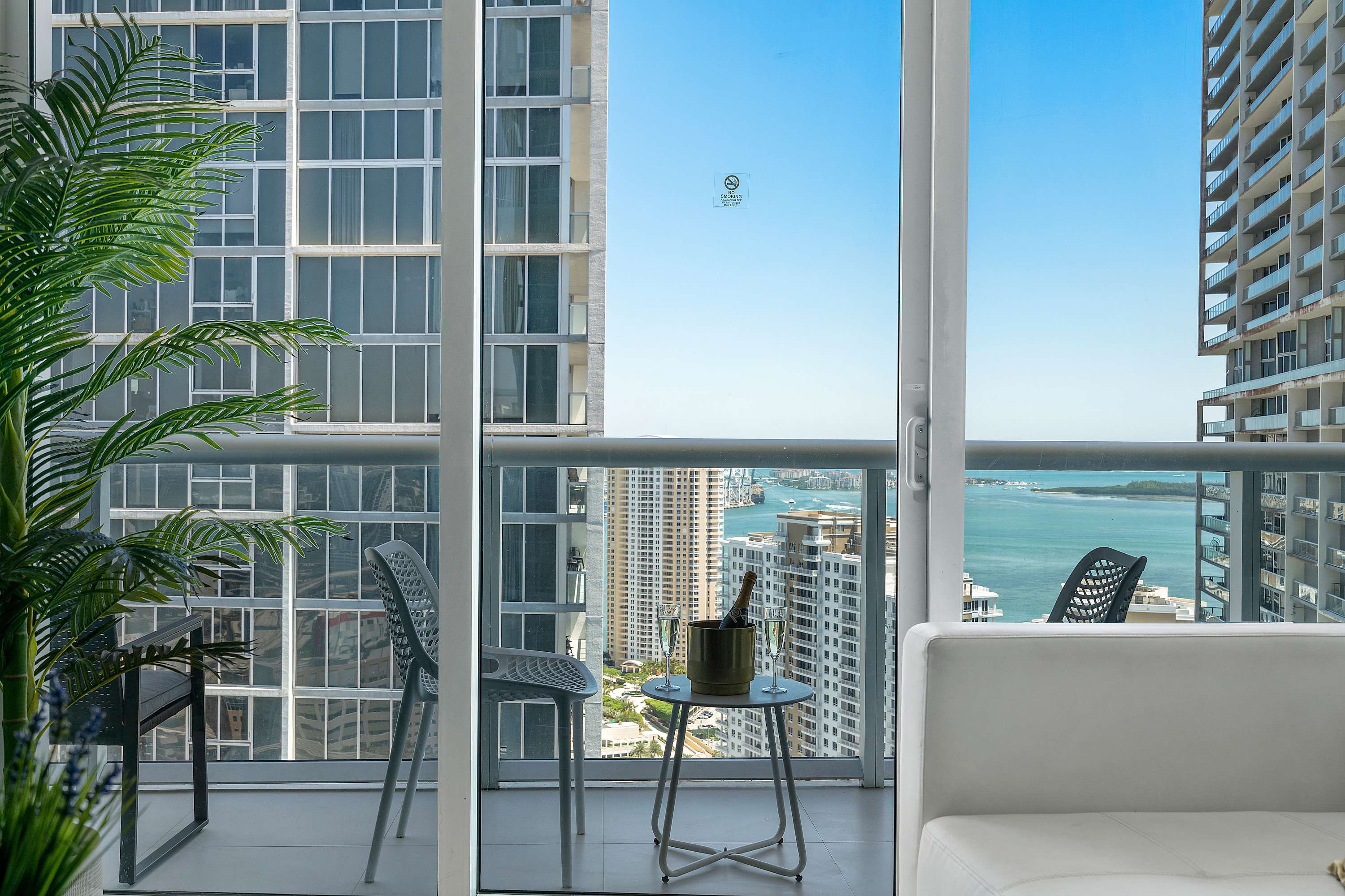 View from the living area looking out onto a balcony with outdoor seating and a champagne bucket, framed by floor-to-ceiling windows showcasing city and water views.