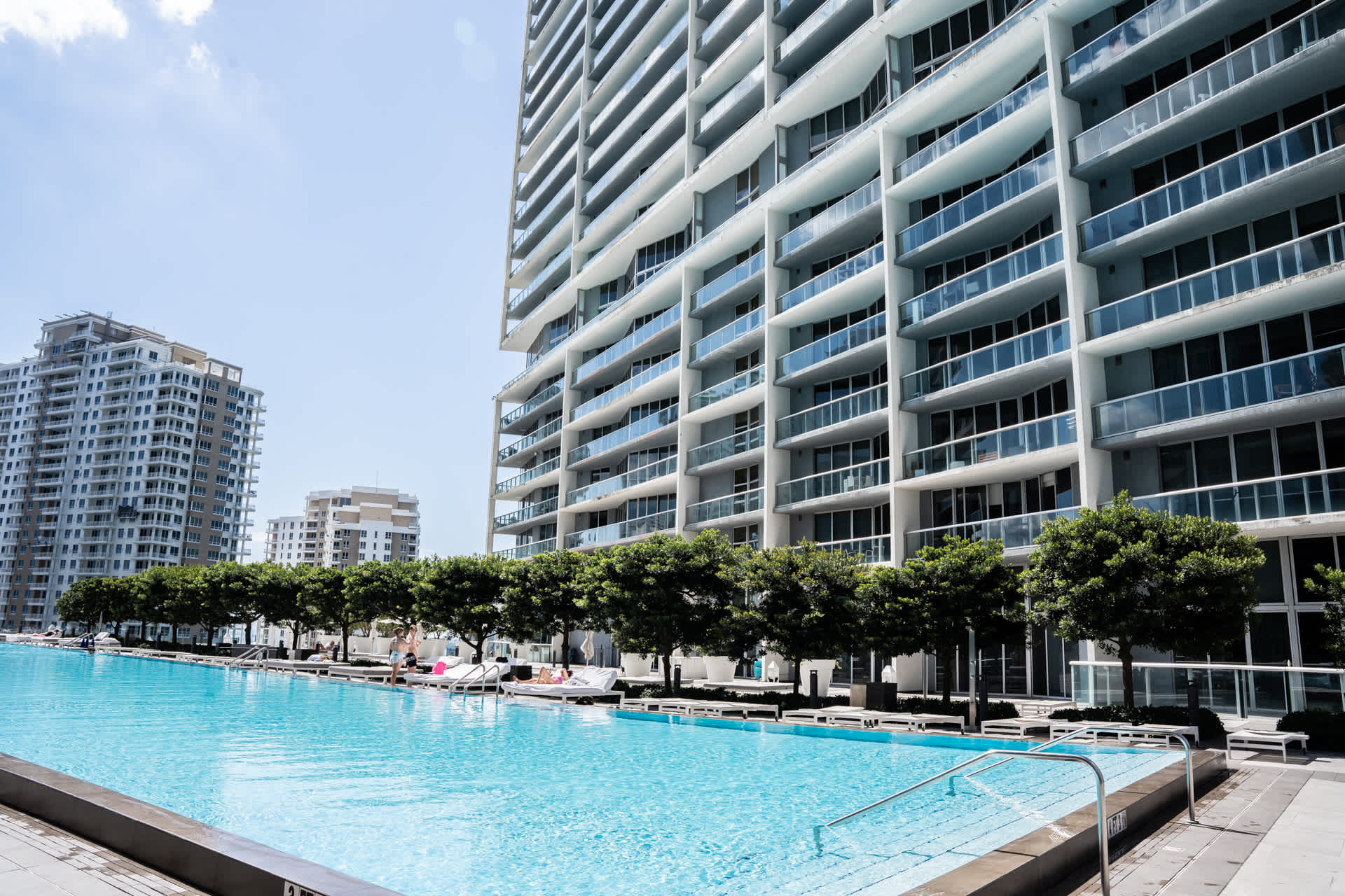 A large, inviting outdoor swimming pool with clear blue water is bordered by a row of green trees and white lounge chairs, with a towering modern high-rise building featuring many balconies dominating the background.