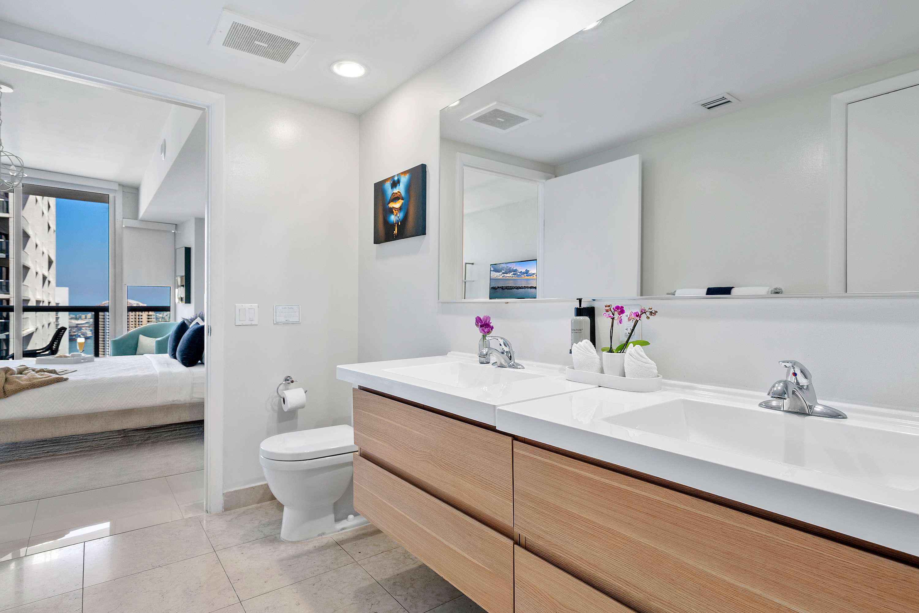 Bathroom showcasing a double vanity with modern fixtures, a toilet, and an open doorway providing a glimpse into the adjacent bedroom, highlighting the clean and contemporary design.