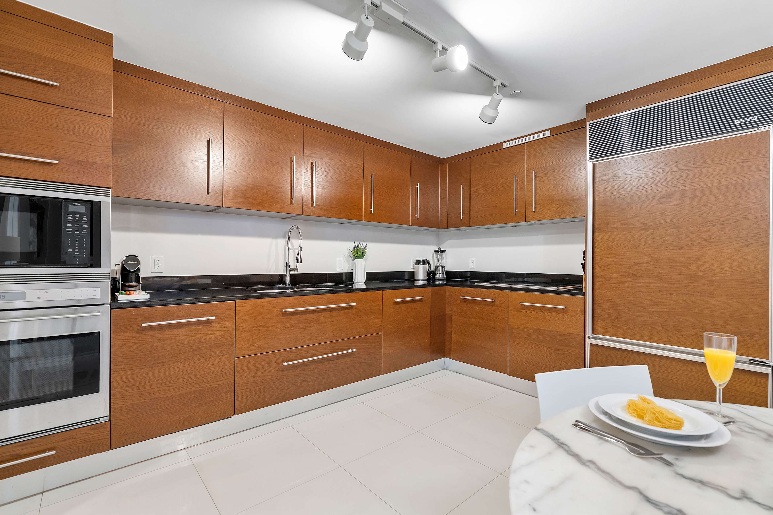 Modern kitchen equipped with dark wood cabinetry, sleek black countertops, stainless steel built-in appliances including an oven and microwave, and track lighting.