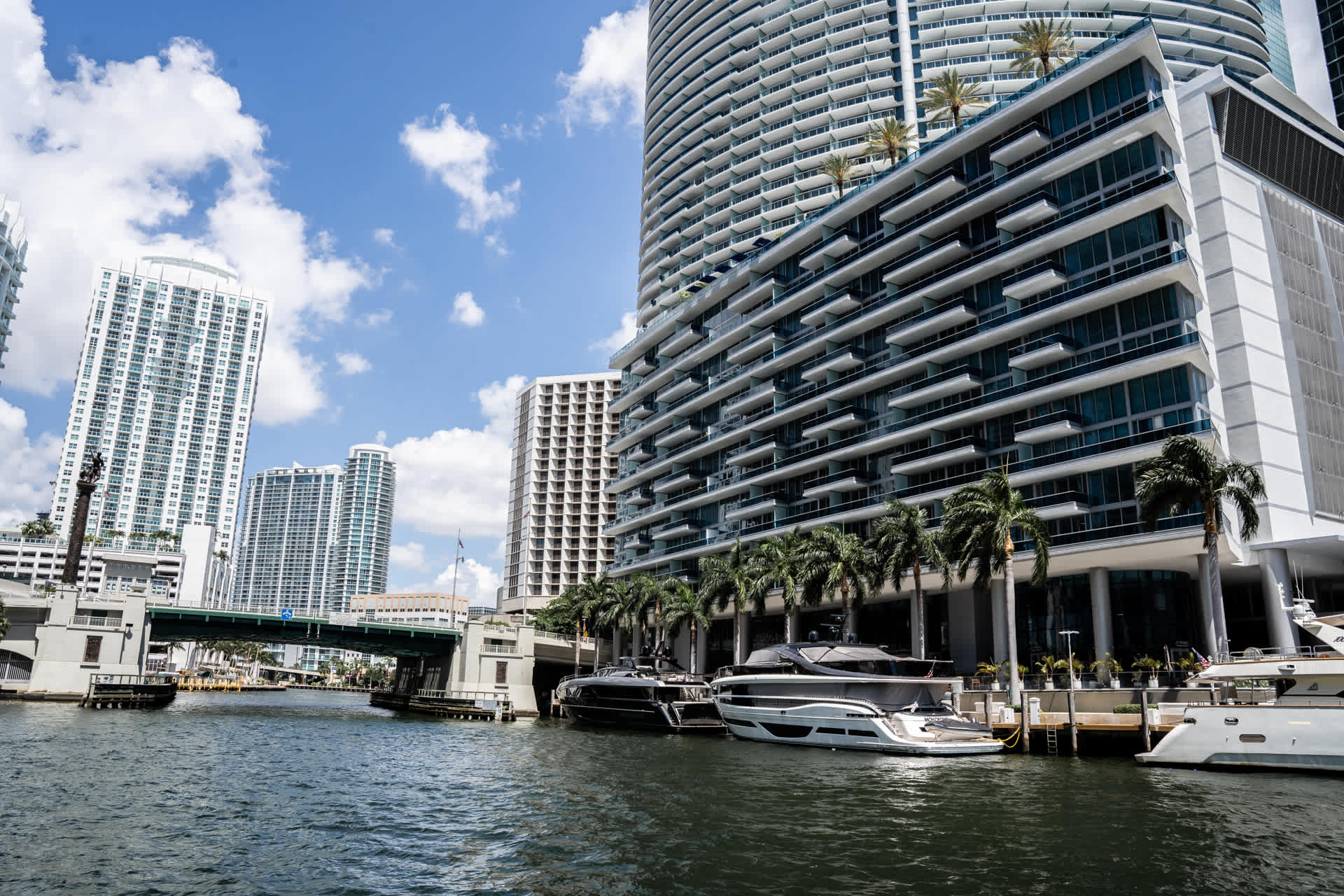 A vibrant exterior view of a modern high-rise building situated along a calm waterway, with several luxury yachts docked nearby. Palm trees line the waterfront, and a bridge is visible in the distance under a clear blue sky.