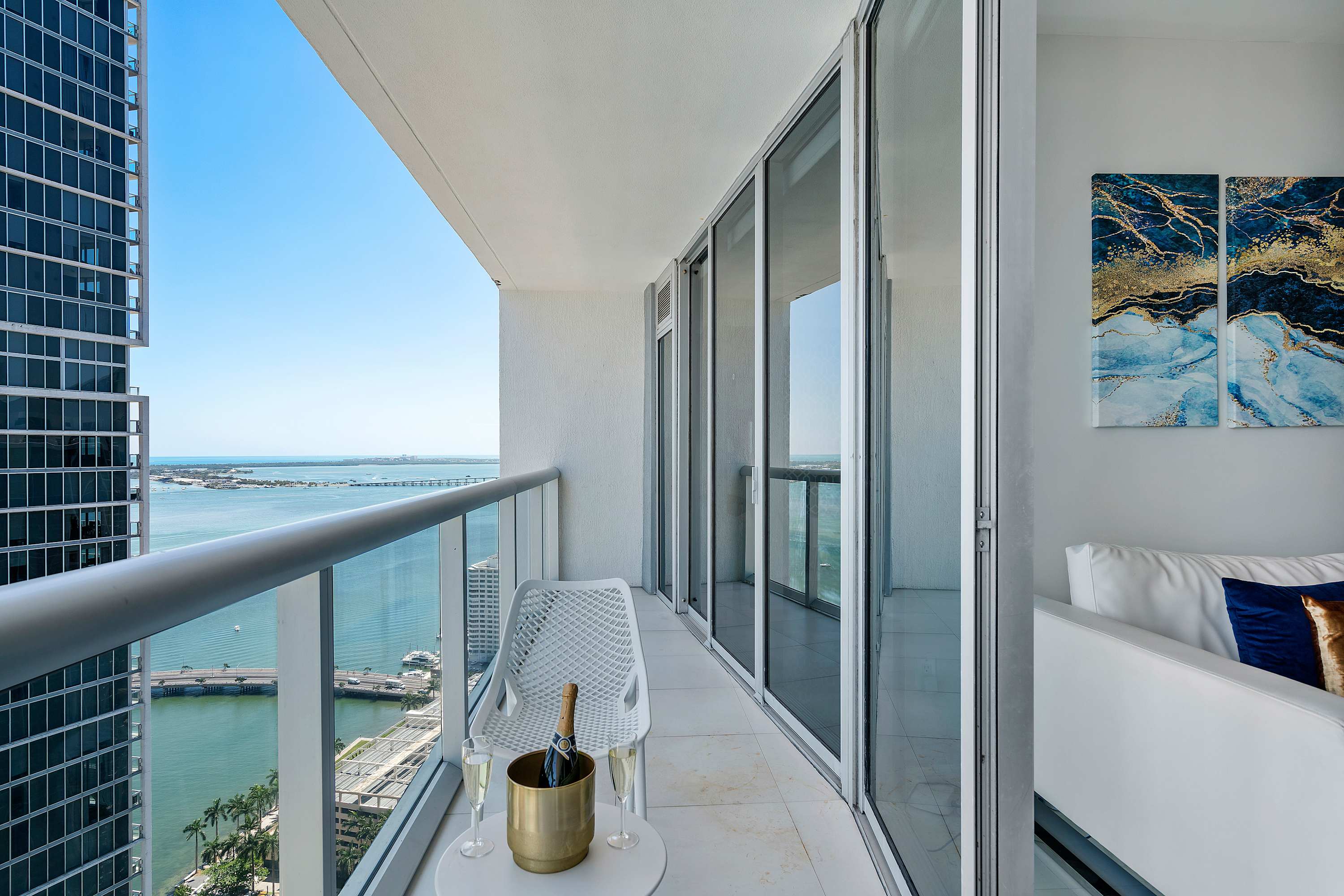 High-rise balcony with a small white outdoor table, a white chair, and a champagne bucket, offering expansive panoramic views of the city skyline and bay.