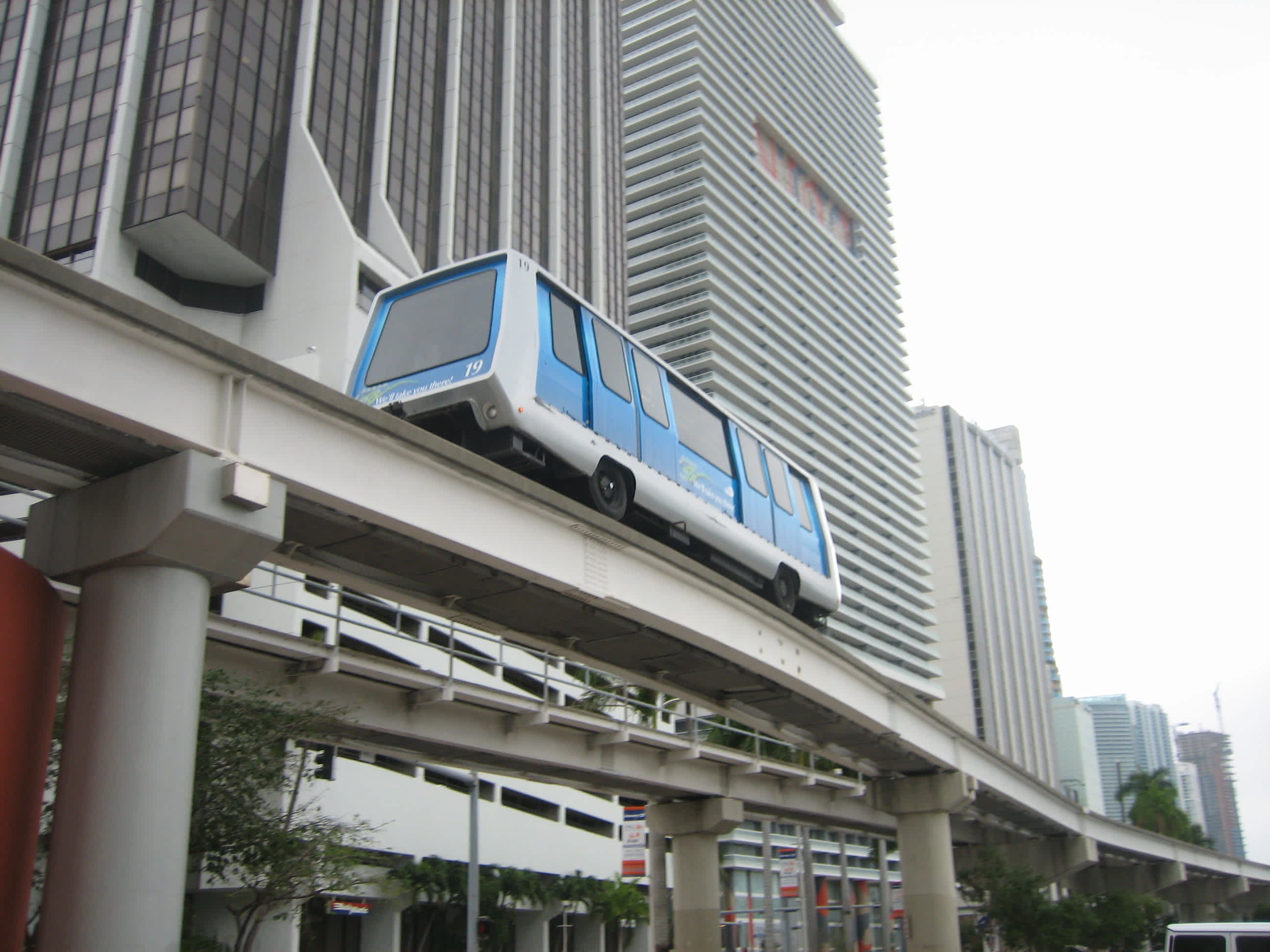 A blue and white monorail train travels on an elevated track through a city environment, with modern high-rise buildings visible in the background under an overcast sky.