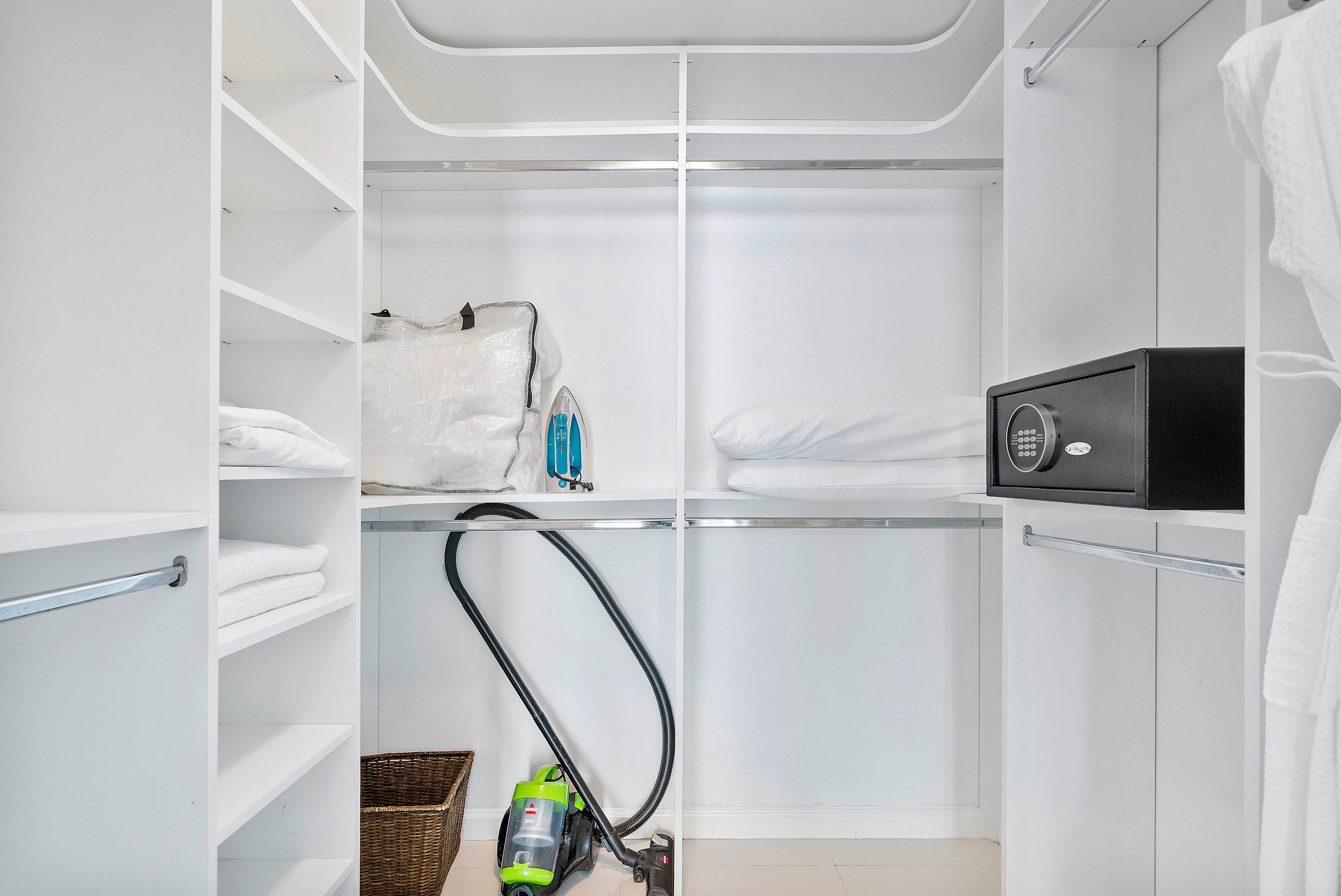 A bright white walk-in closet features multiple shelves stocked with neatly folded white linens, a black digital safe, an iron, and a green and black vacuum cleaner stored on the floor.