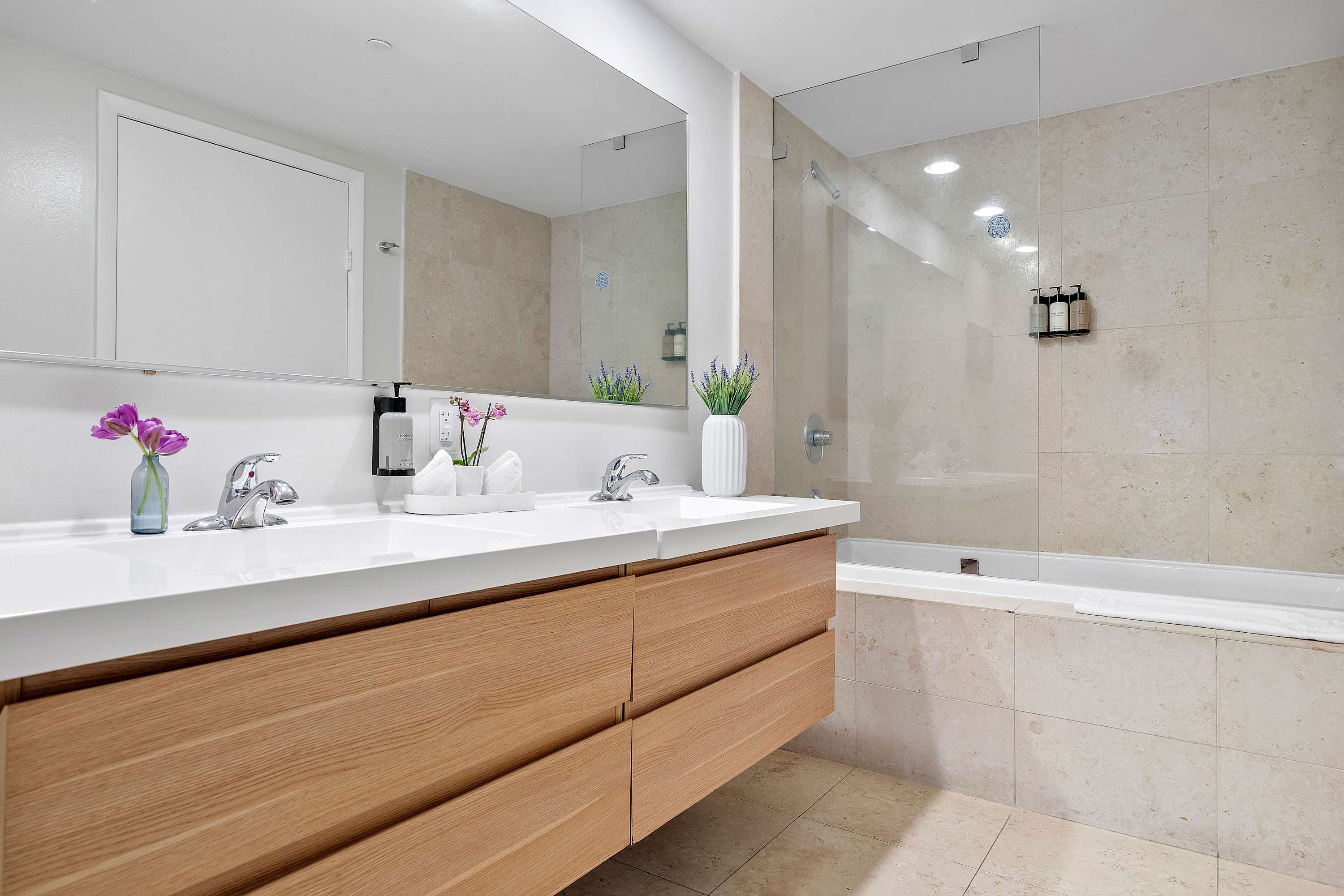 Modern bathroom with a double vanity featuring light wood cabinets and white countertops, a large mirror, and a combination shower-tub with beige tiled walls.
