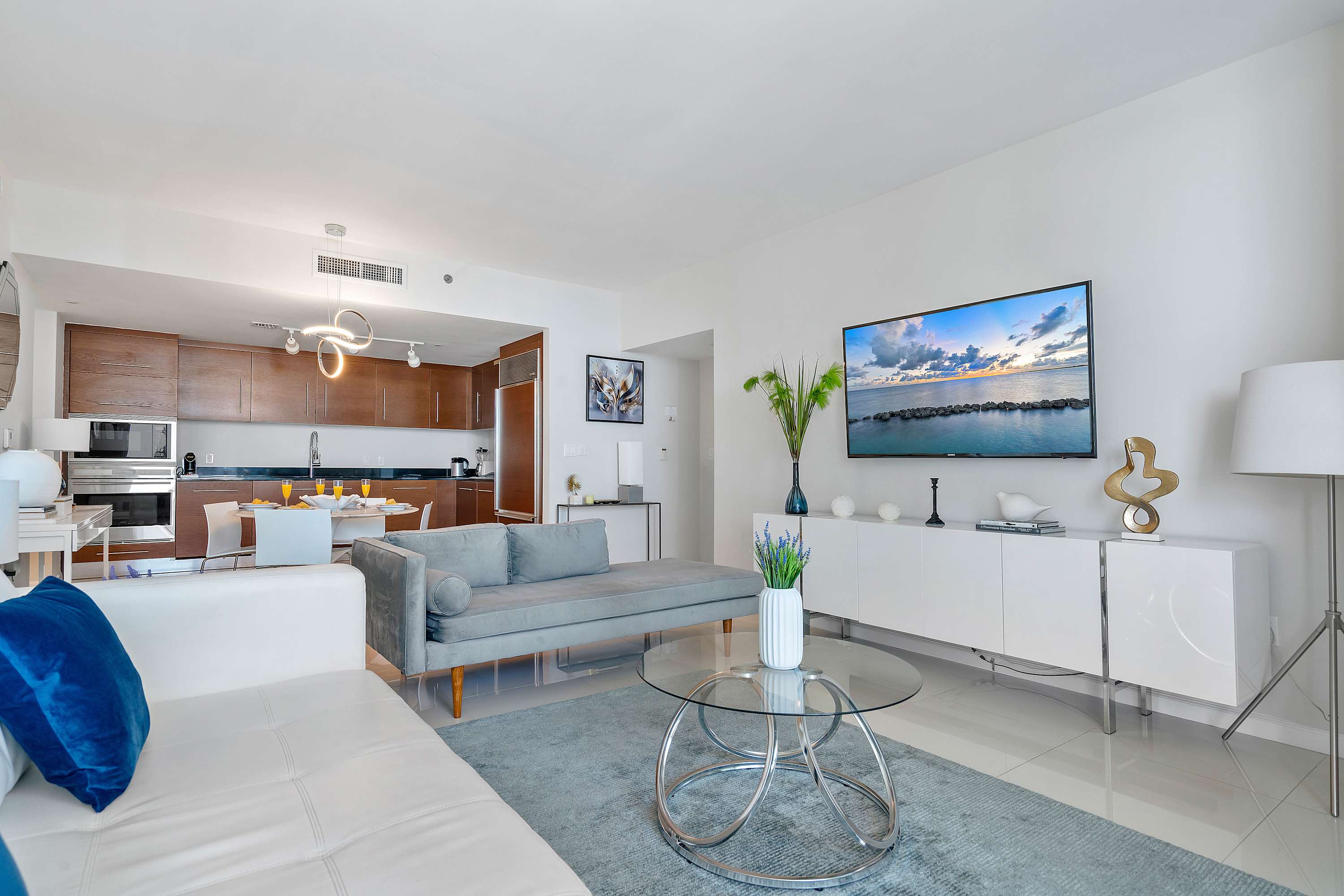 Contemporary living room with a white sofa, grey chaise lounge, glass coffee table, and a large wall-mounted TV, seamlessly connected to the kitchen and dining areas.