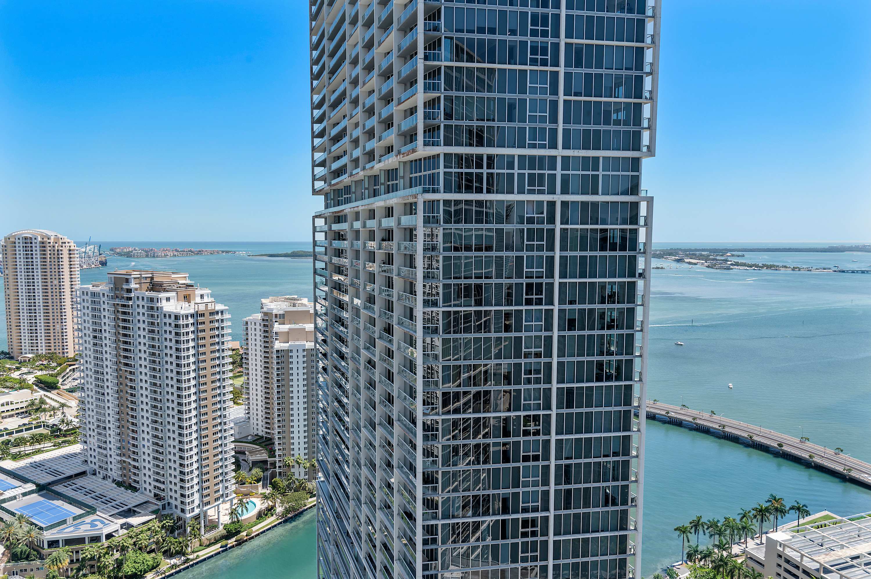 A high-angle view captures a towering modern skyscraper with a glass facade and numerous balconies, set against a backdrop of a vibrant city, expansive blue waters, and distant bridges under a clear sky.