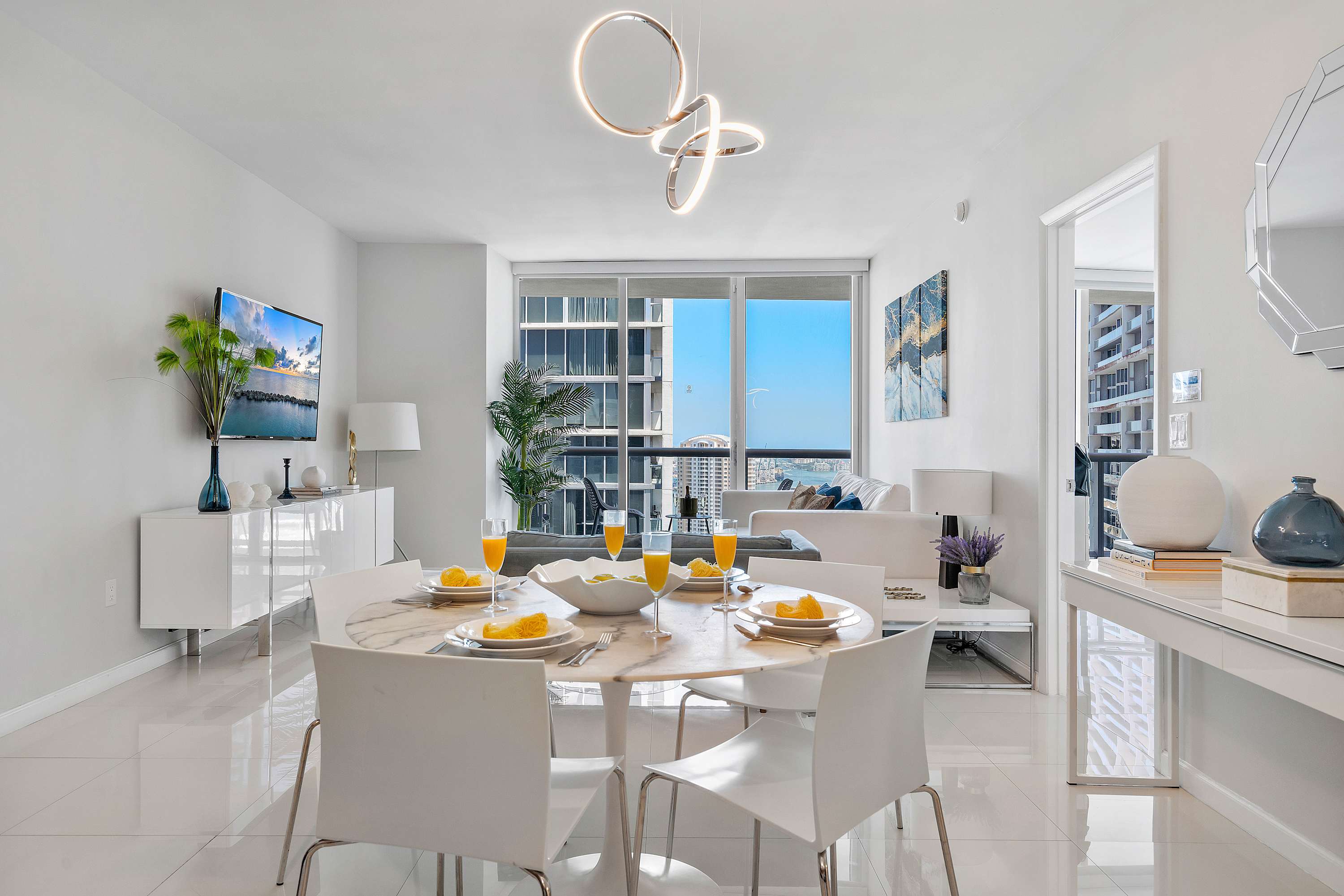 Open-concept dining area featuring a round marble-top table with white chairs, illuminated by a modern circular chandelier, adjacent to a living space and balcony with city views.