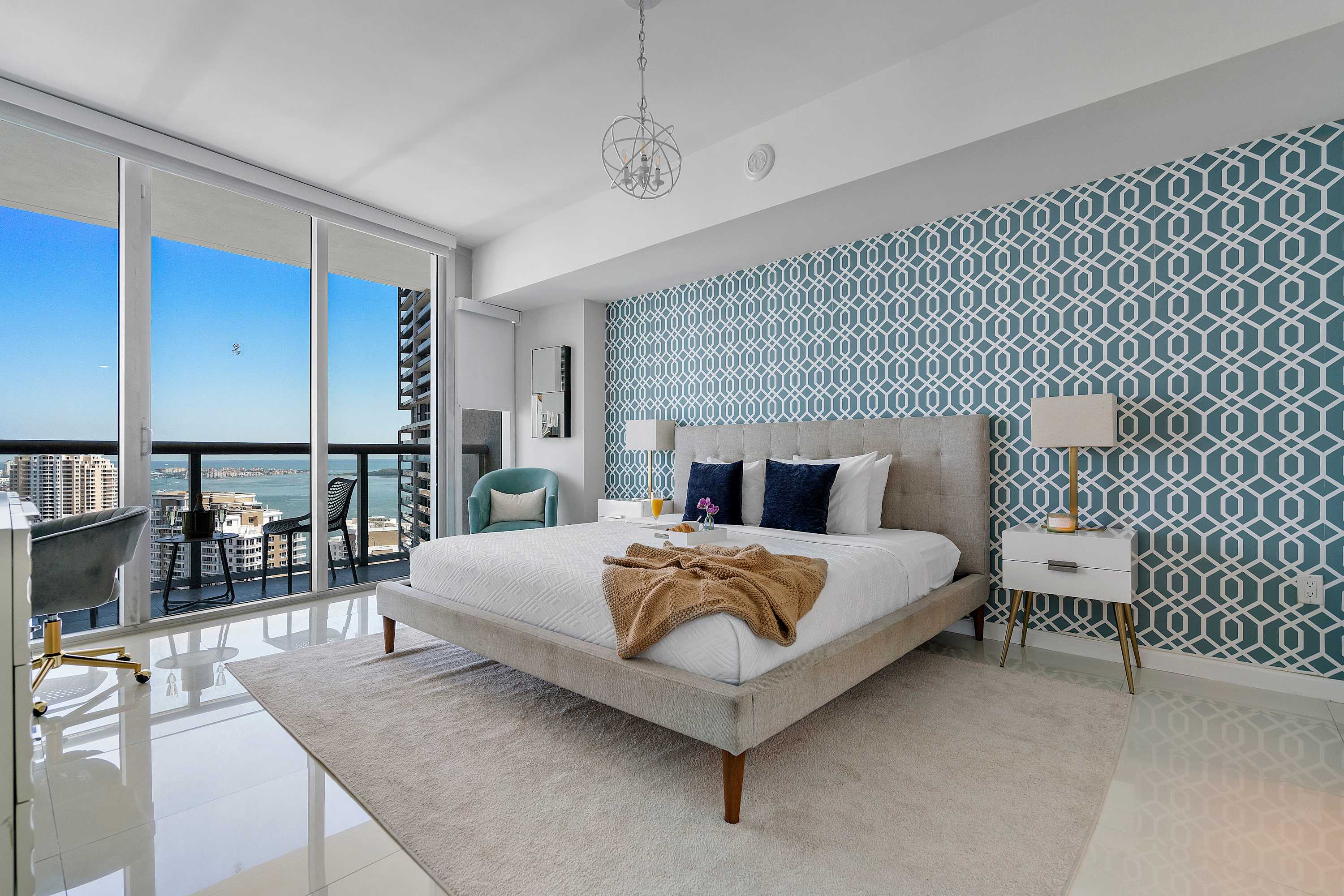 Bright bedroom with a large bed, grey upholstered headboard, blue geometric accent wall, white nightstands, and a wide window showcasing city and water views.