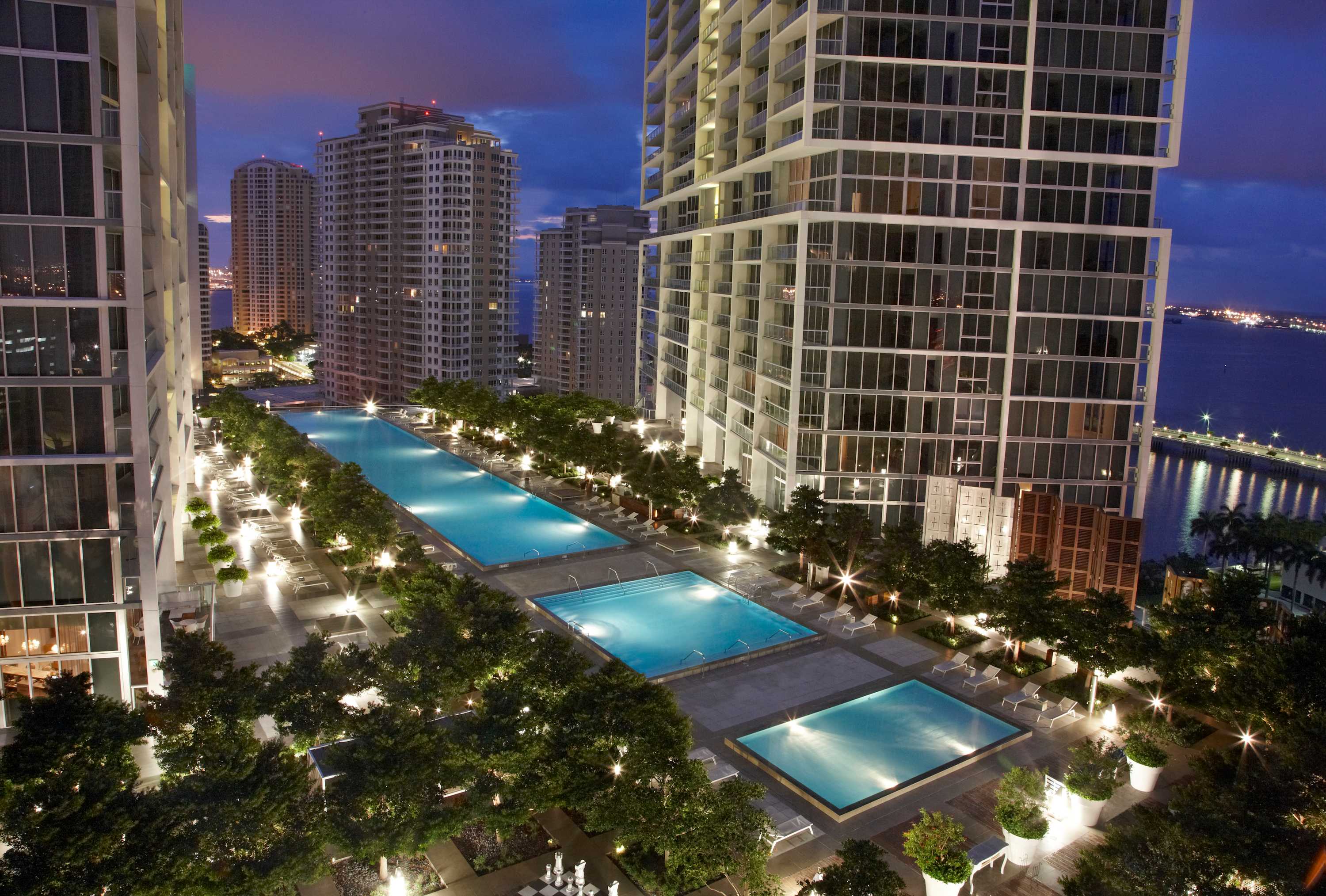 A stunning aerial view of a large outdoor pool deck at dusk, featuring multiple illuminated swimming pools surrounded by lush green trees and lounge chairs. High-rise buildings tower around the deck, with city lights and a waterfront visible in the distance.