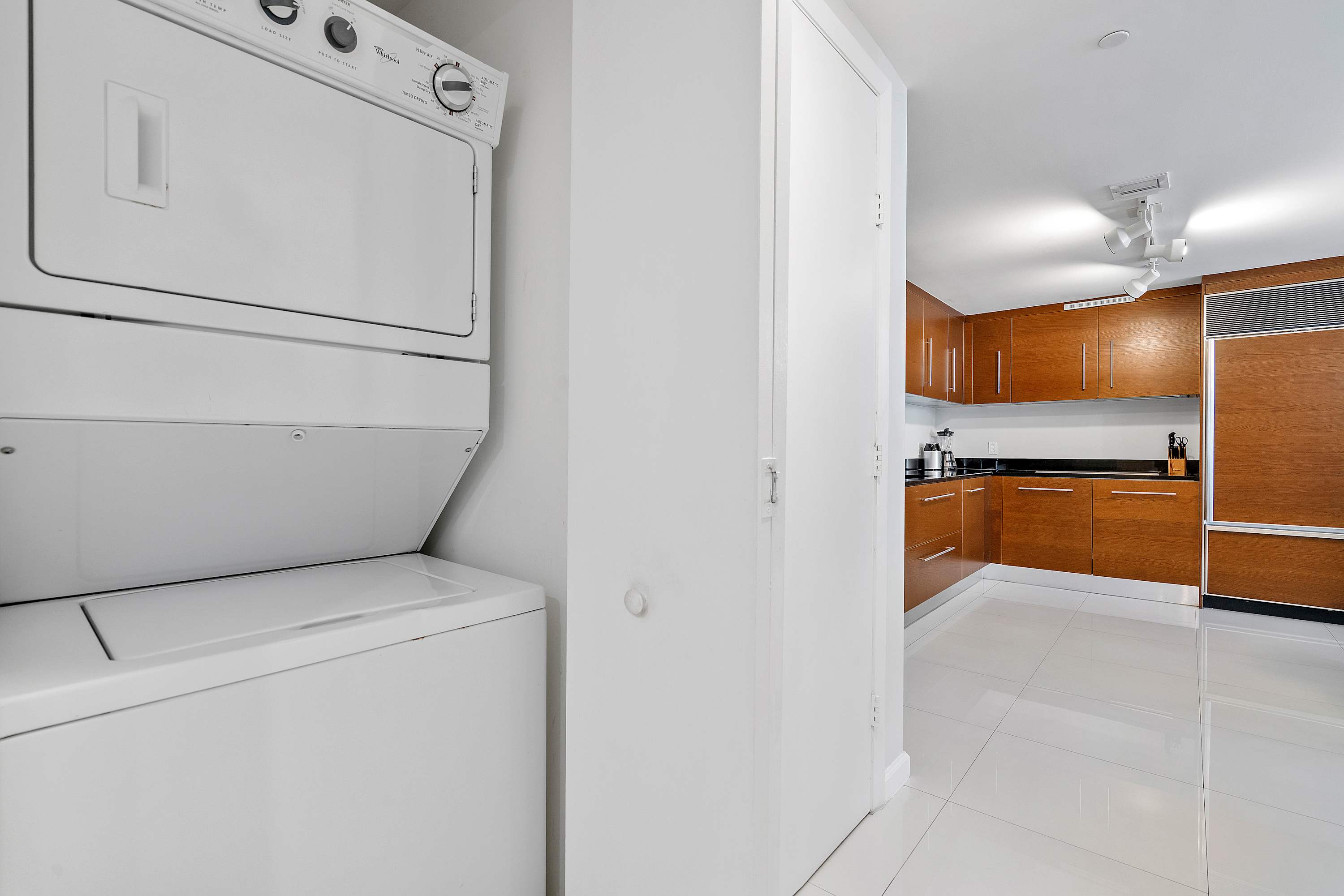 A white stacked washer and dryer unit is neatly placed in a bright utility area, with a glimpse of a modern kitchen featuring warm wooden cabinetry and sleek white tiled floors in the background.