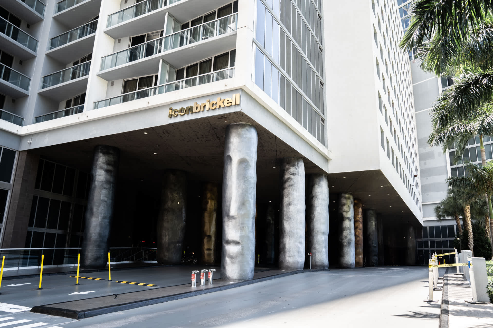 The grand entrance of a modern high-rise building, featuring a covered driveway supported by large, sculptural columns with carved faces. The building's name, "ICONBRICKELL," is prominently displayed above the entrance.