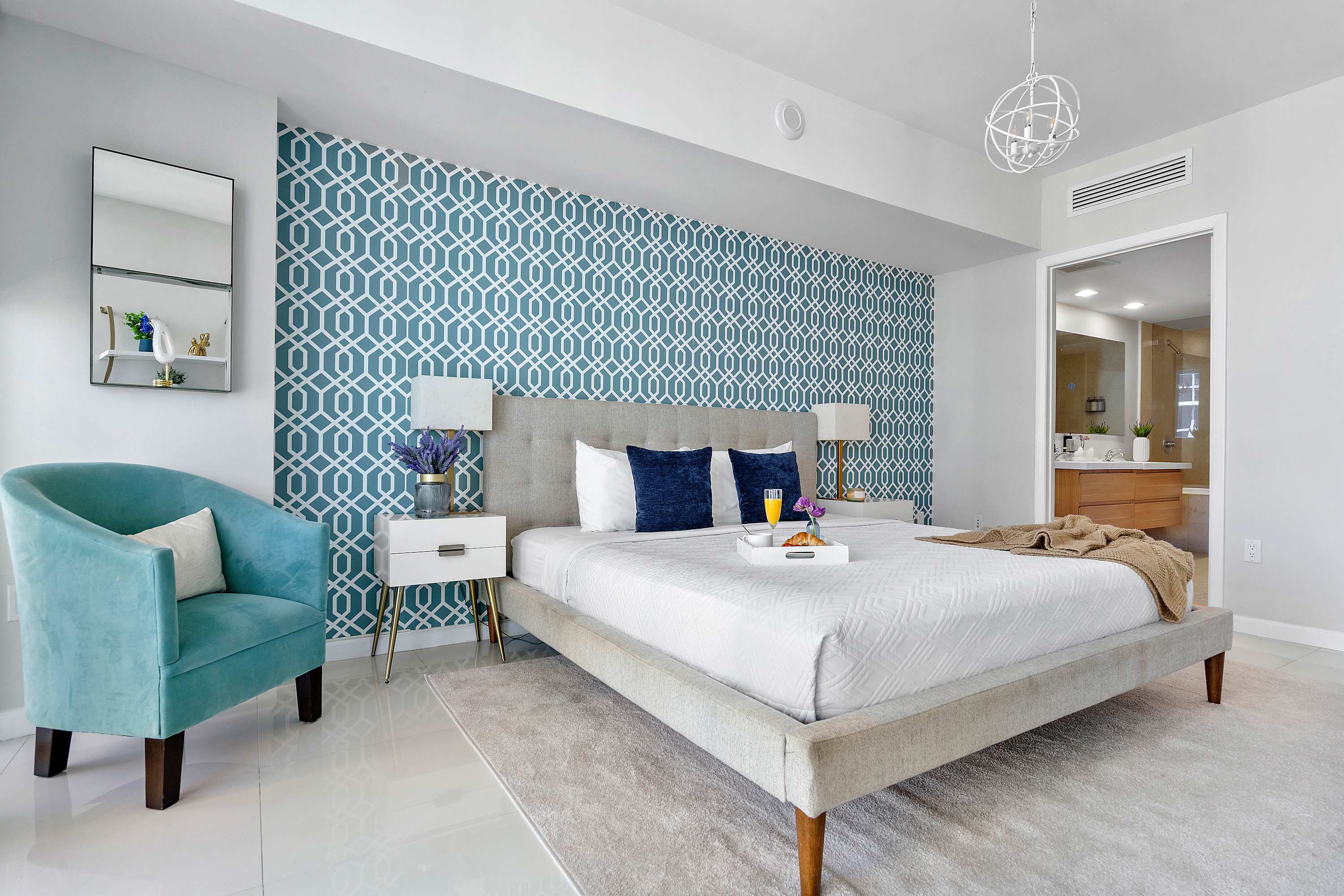 Master bedroom featuring a grey upholstered bed, a striking blue geometric accent wall, white nightstands, a light blue accent chair, and an open doorway leading to the bathroom.