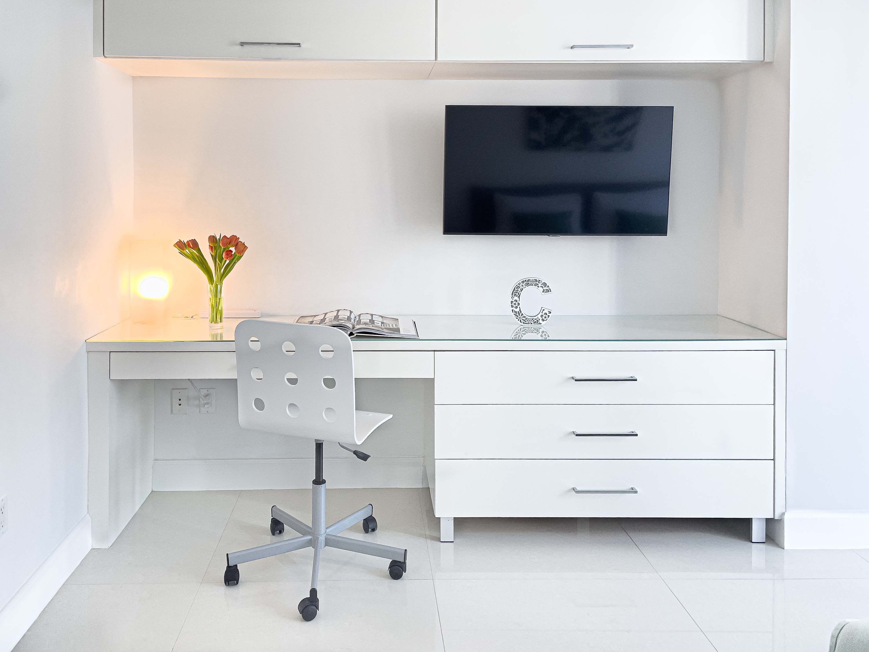 Dedicated workspace with a white desk, matching drawers, and a white rolling office chair. A wall-mounted television is above the desk, and a vase with flowers adds a touch of color.
