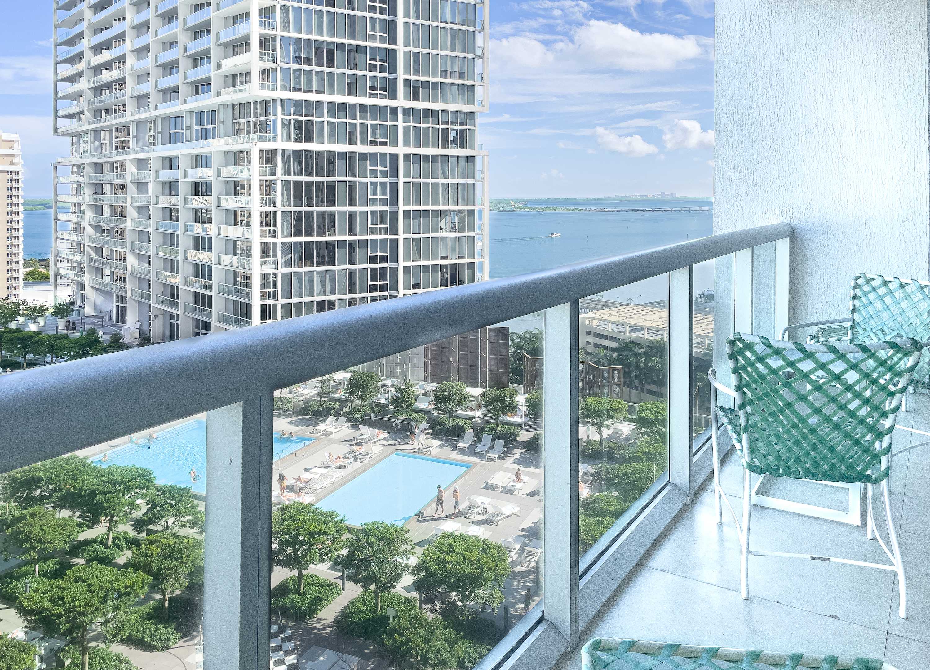 A high-rise balcony with a glass railing offers a panoramic view of a city skyline, a large outdoor pool area with lush trees, and a distant bay with boats under a partly cloudy sky. Two green woven chairs are visible on the tiled balcony floor.
