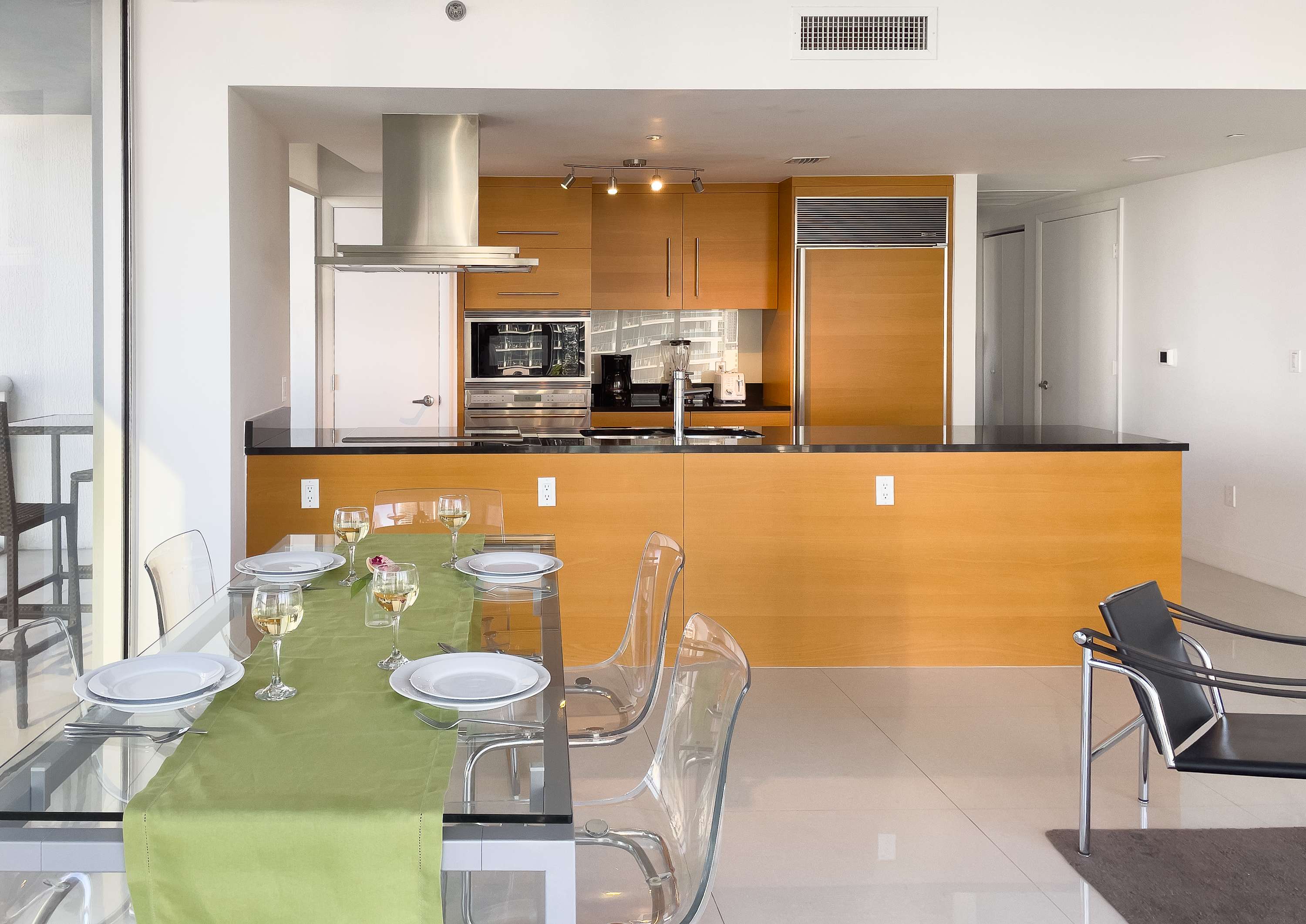 Modern kitchen with light wood cabinetry, black countertops, and stainless steel appliances, including a range hood and built-in oven. Adjacent is a glass dining table set with clear chairs and place settings.