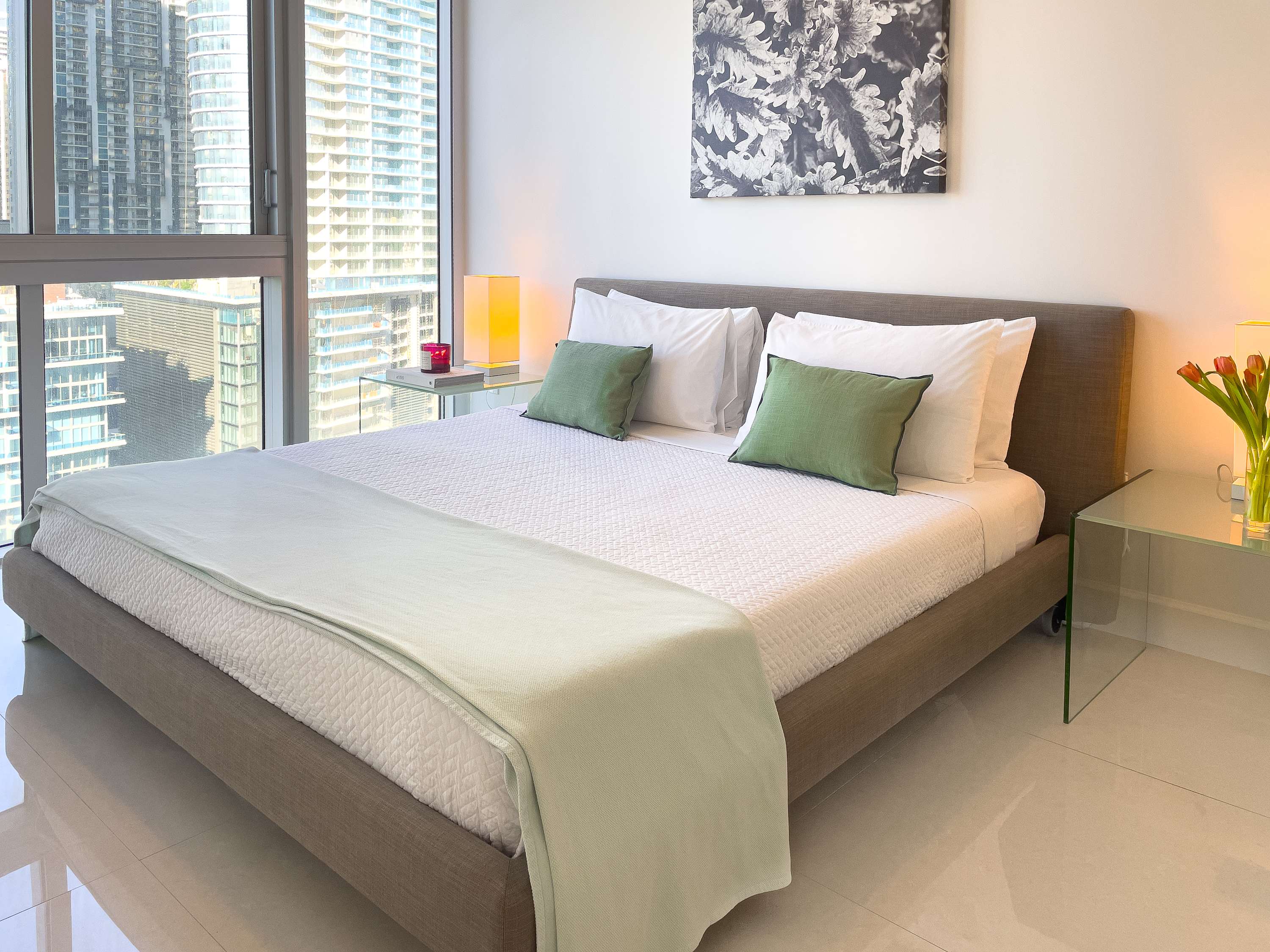 Bright bedroom featuring a queen-sized bed with a light grey frame, white bedding, and green accent pillows. Floor-to-ceiling windows provide a city view, and glass nightstands flank the bed.
