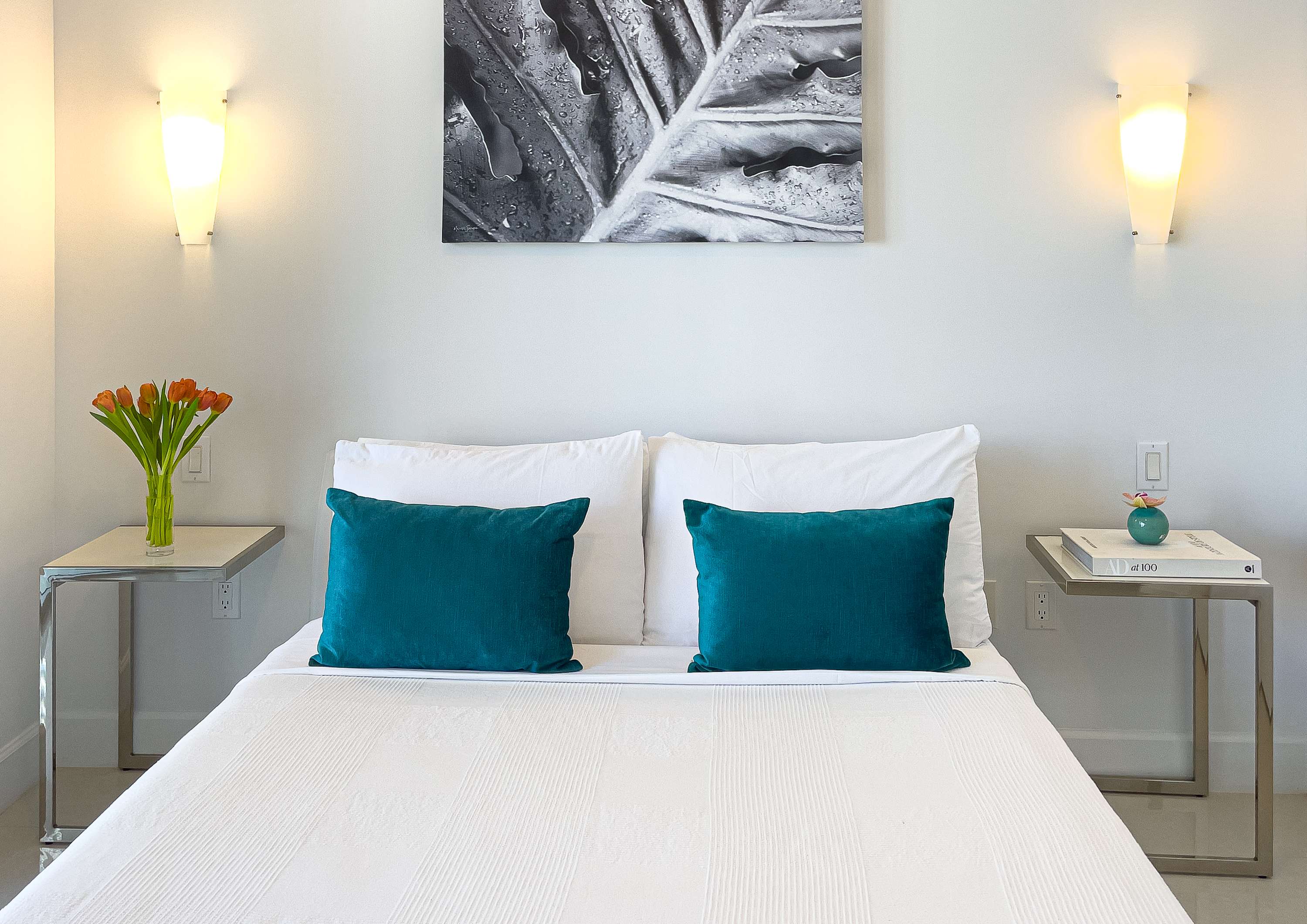 Cozy bedroom featuring a queen-sized bed with white bedding and two teal accent pillows. Two modern metal nightstands with glass tops flank the bed, each with a lamp and decorative items.