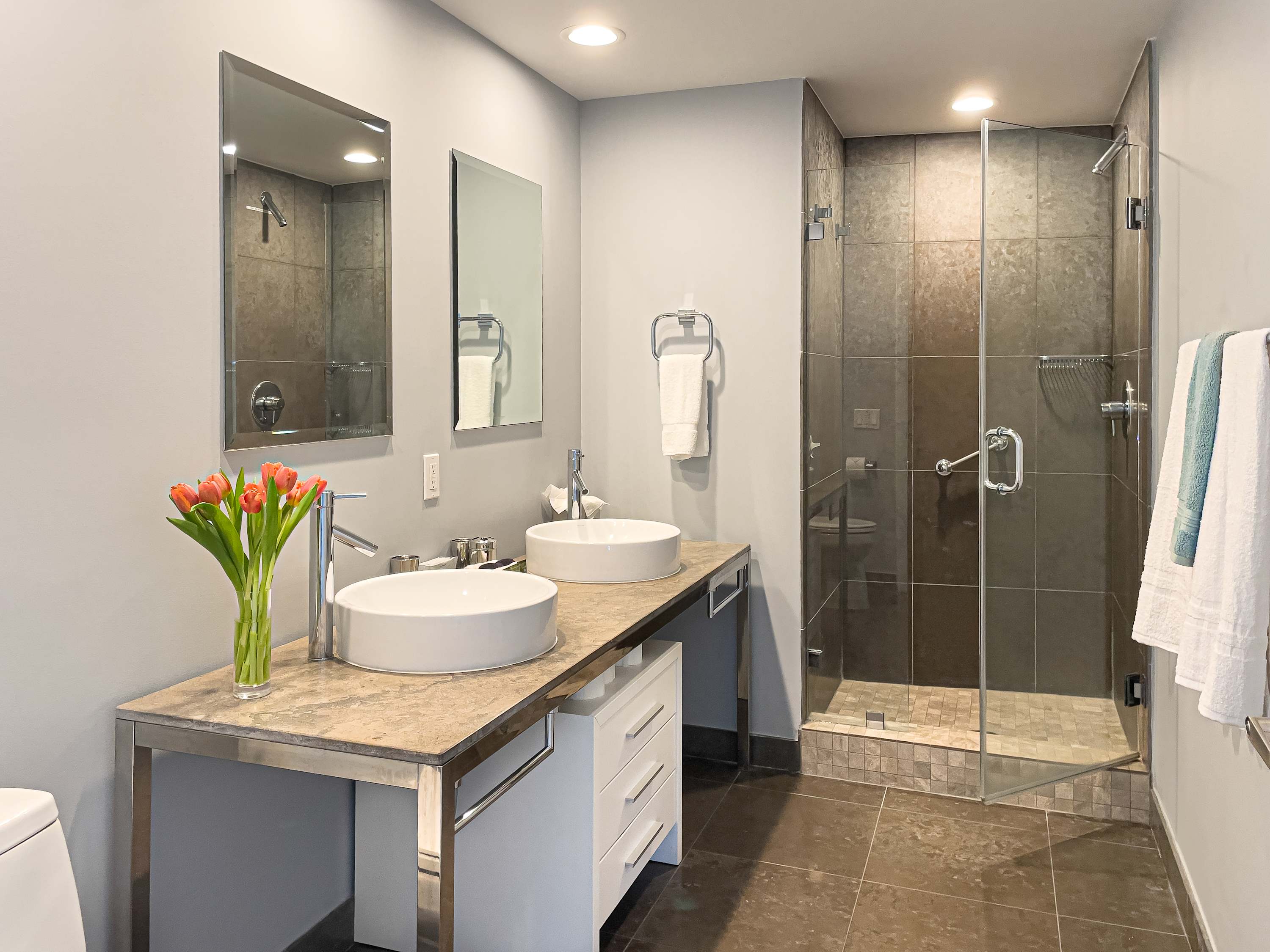 Contemporary bathroom with a double vanity featuring two vessel sinks on a stone countertop with a metal frame. A glass-enclosed walk-in shower is visible to the right, and a large mirror hangs above the sinks.