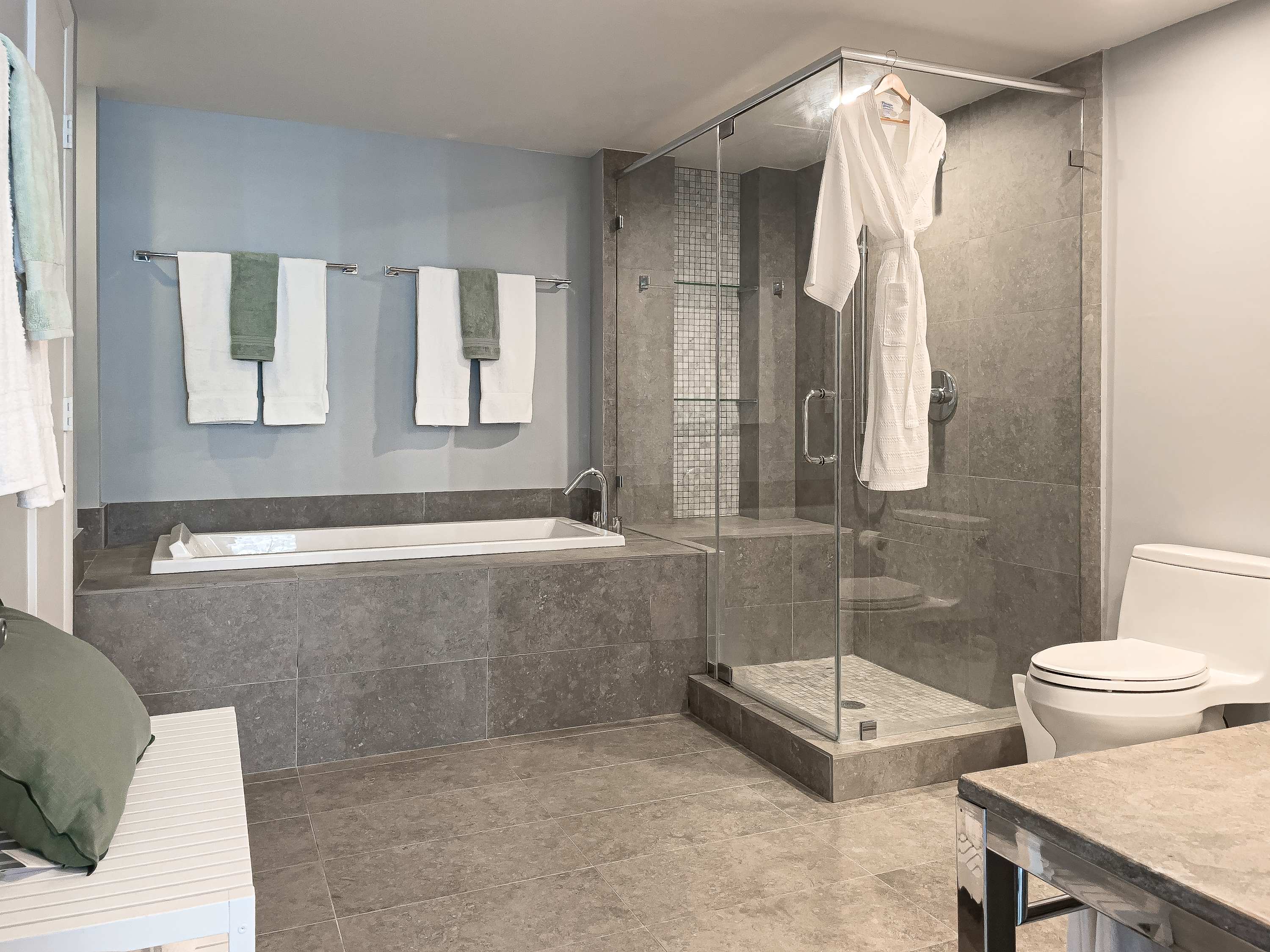 Spacious modern bathroom with grey tiled floors and walls. It features a built-in bathtub and a large glass-enclosed walk-in shower with a tiled accent wall.