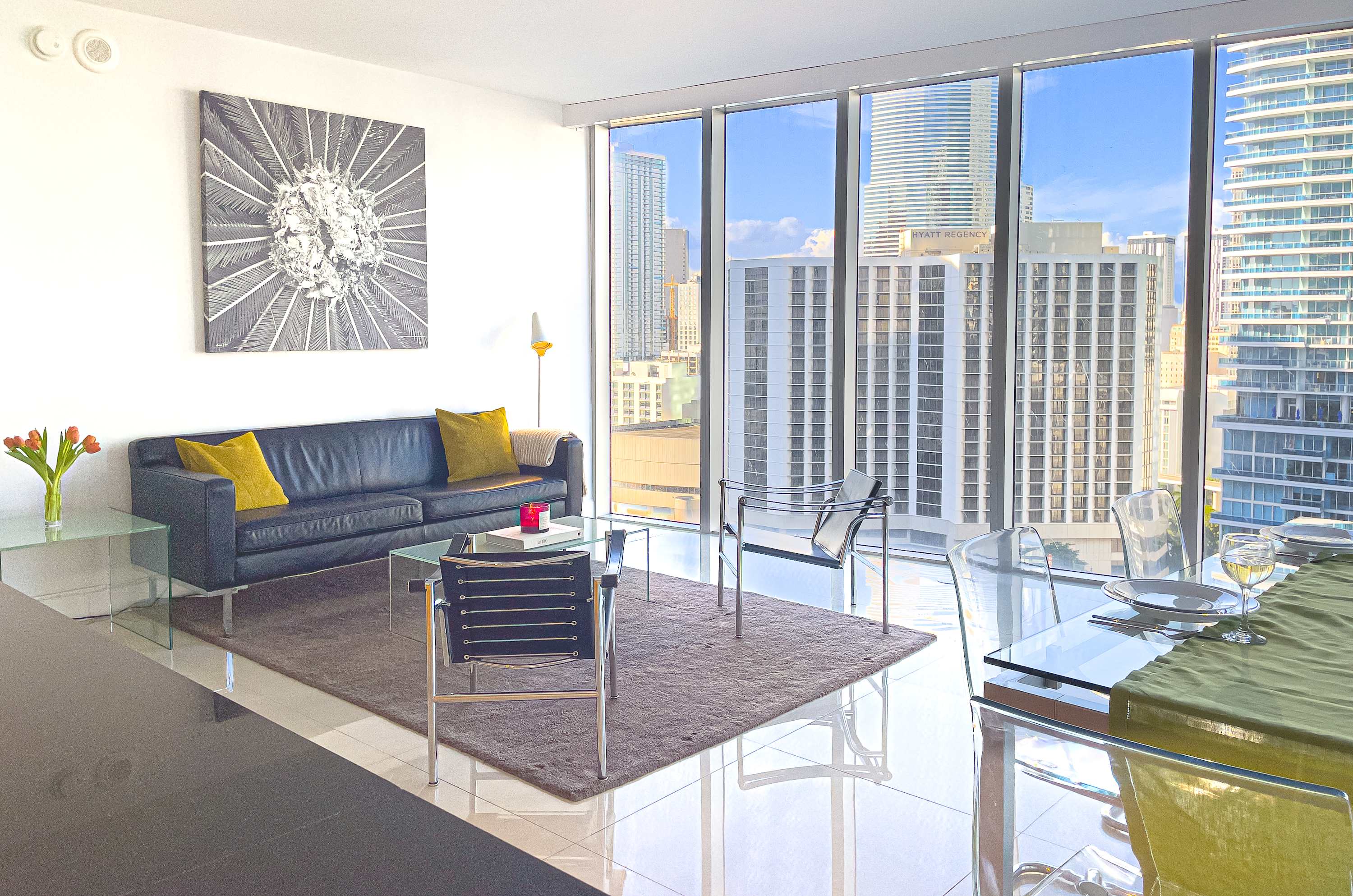 Open-concept living and dining area with a black leather sofa, glass coffee table, and two black metal chairs on a dark rug. A glass dining table with clear chairs is visible, all set against a backdrop of floor-to-ceiling windows with city views.