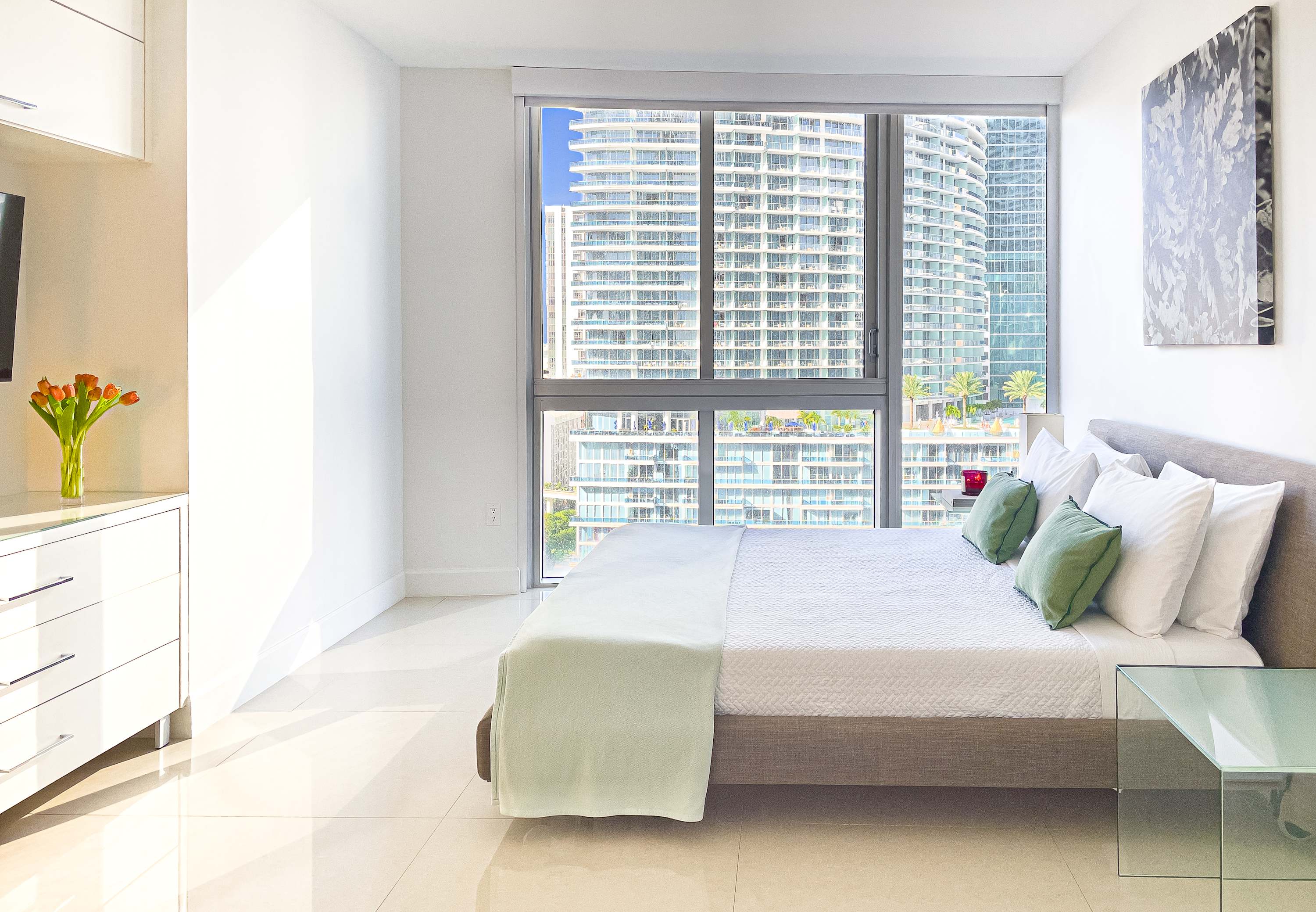 Bright bedroom with a queen-sized bed, white bedding, and green accent pillows. Large windows offer a view of surrounding high-rise buildings, and a white dresser with a vase of flowers is visible on the left.