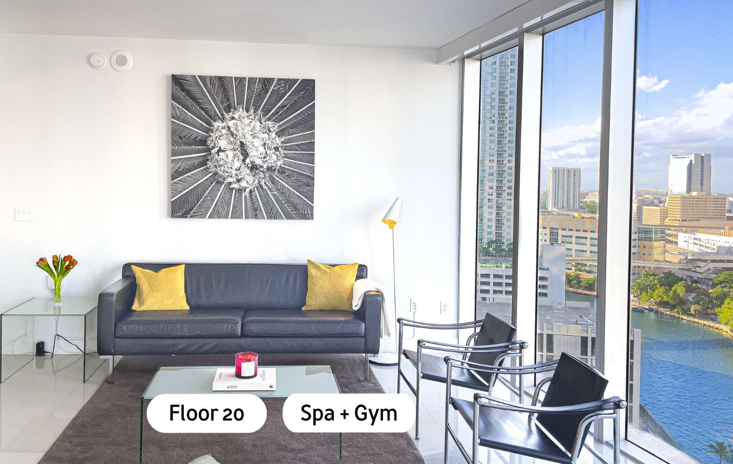 Modern living room featuring a black leather sofa with yellow accent pillows, a glass coffee table, and two black metal chairs, all set on a dark rug. Floor-to-ceiling windows offer panoramic city and waterfront views.