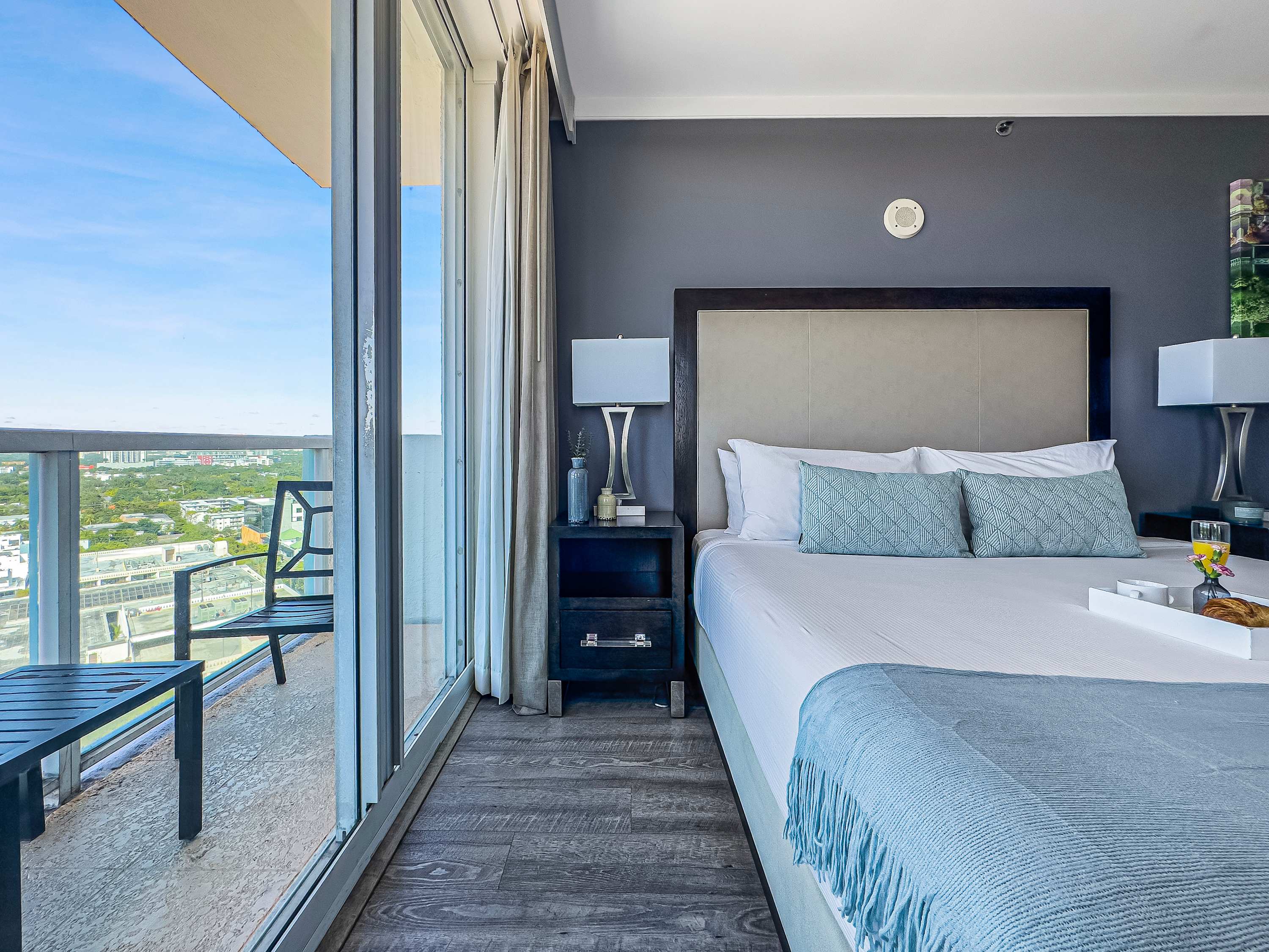 A king-sized bed with light blue bedding is positioned next to a large glass sliding door leading to a balcony with outdoor seating. A dark nightstand with a lamp sits beside the bed, and a city view is visible through the door.