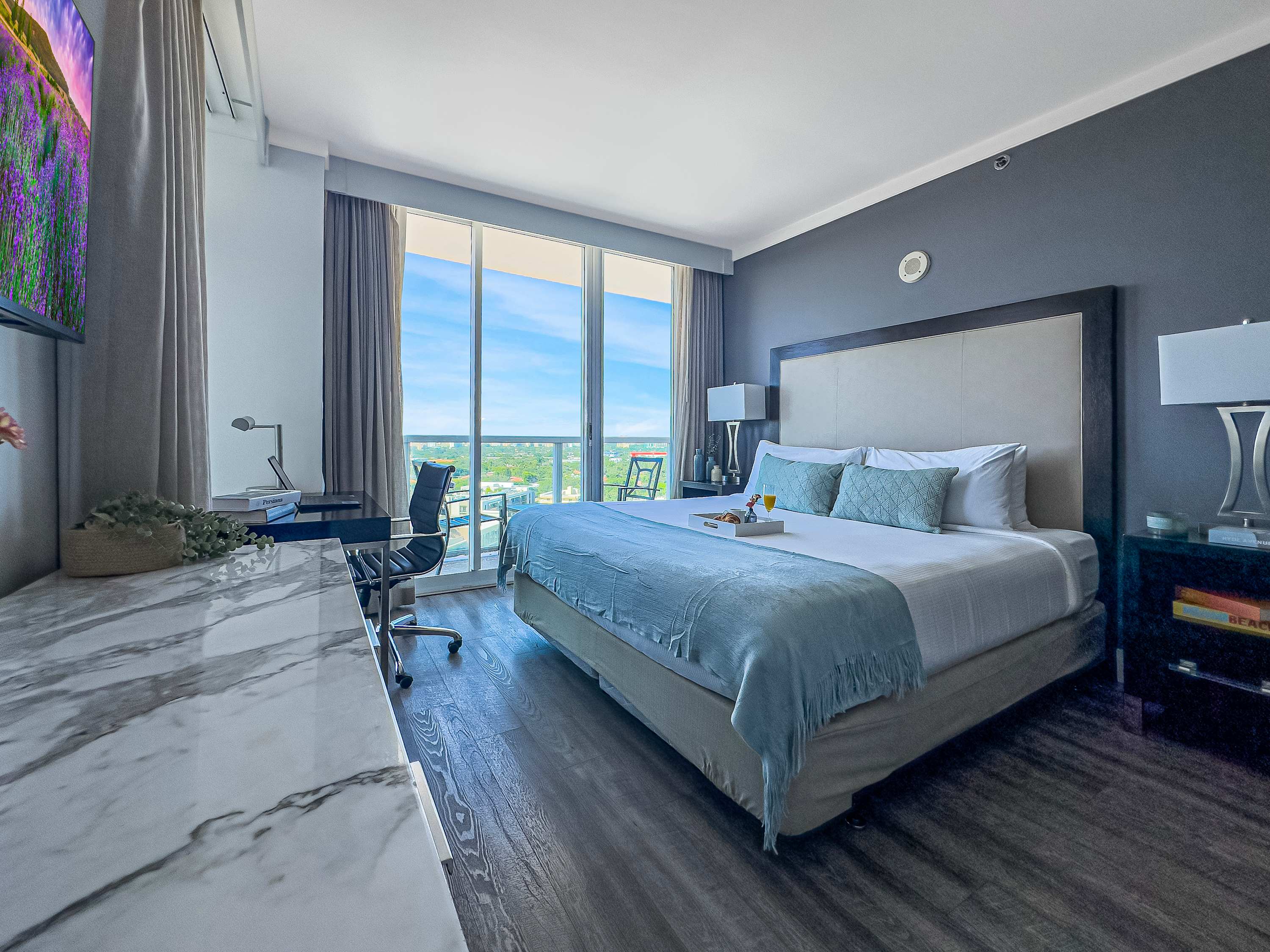 A modern studio bedroom features a king-sized bed with light blue bedding, a dark gray accent wall, and a dedicated workspace with a desk and chair by large windows. The windows offer a panoramic city view with lush greenery.