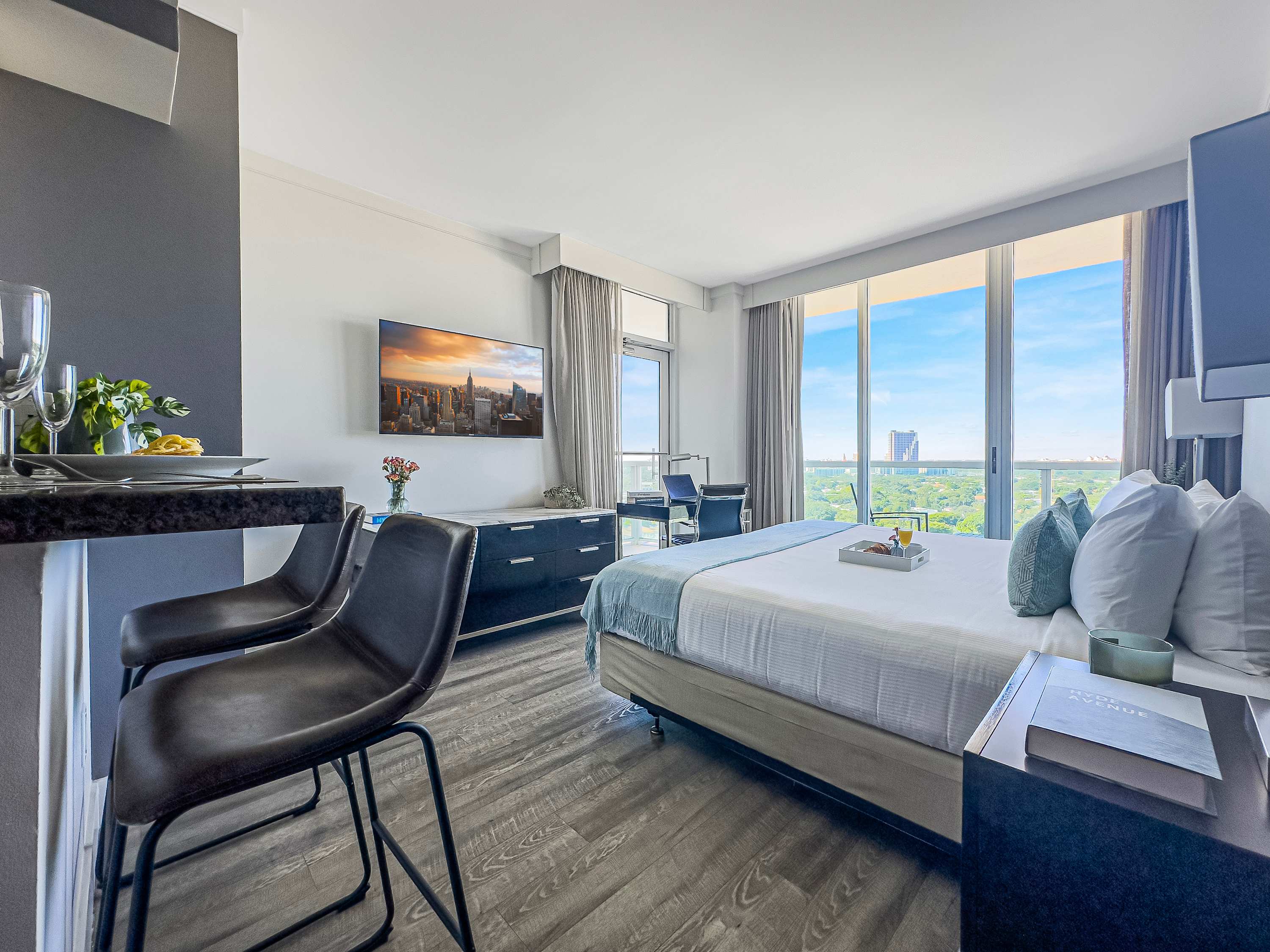 A bright studio bedroom features a king-sized bed with light blue accents, a wall-mounted TV, and a desk with an office chair near large windows offering a city view. A small kitchen bar with two stools is visible on the left.