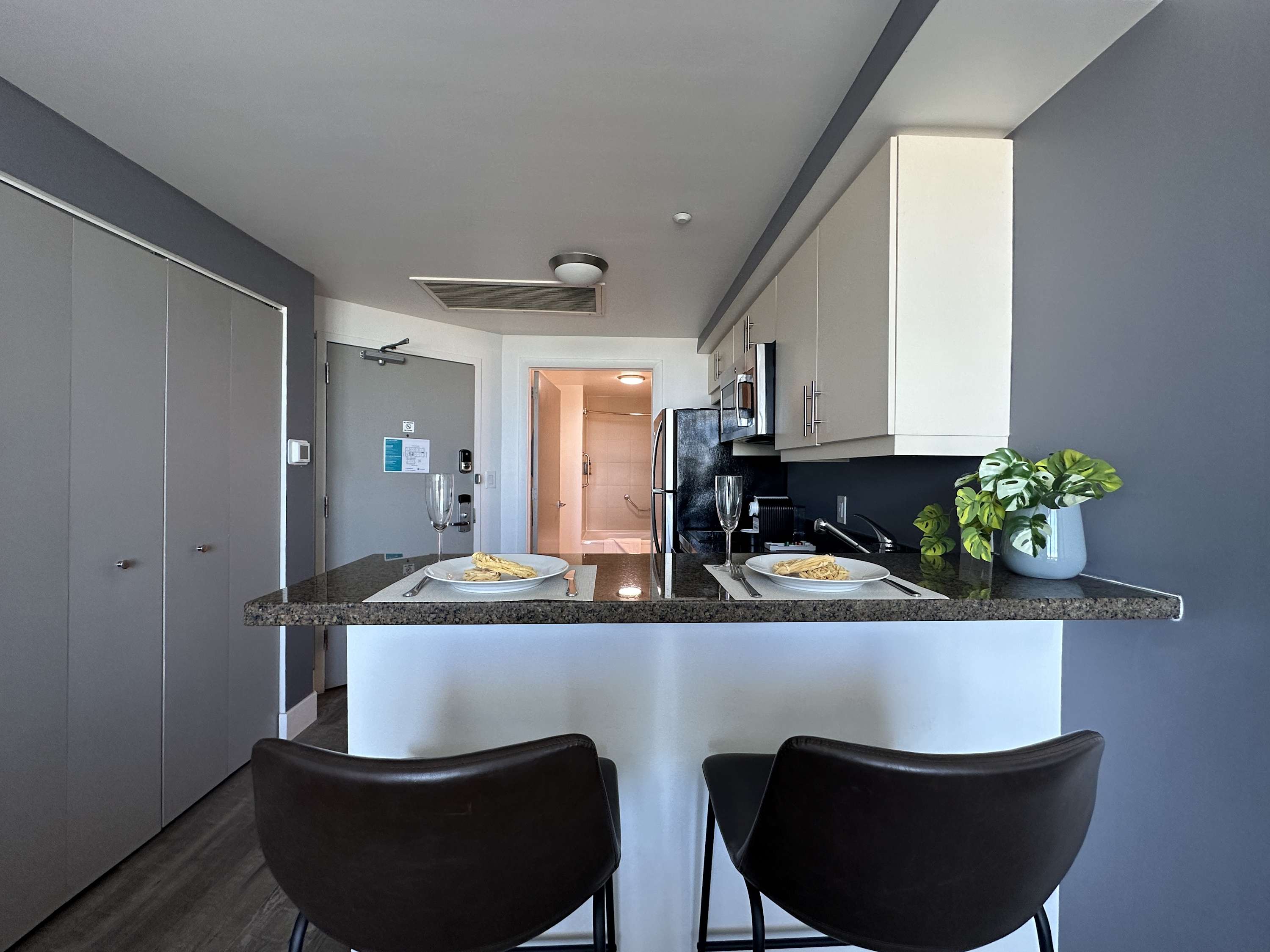 A modern kitchenette bar with two dark stools and place settings for two is positioned against a dark gray wall, facing the entrance to the studio. Light-colored cabinets and a black refrigerator are visible in the background.