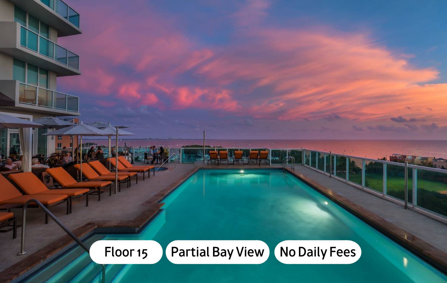 An elevated outdoor swimming pool with orange lounge chairs and umbrellas, overlooking a bay or ocean at sunset with a vibrant pink and purple sky. A modern building with glass balconies is visible on the left.