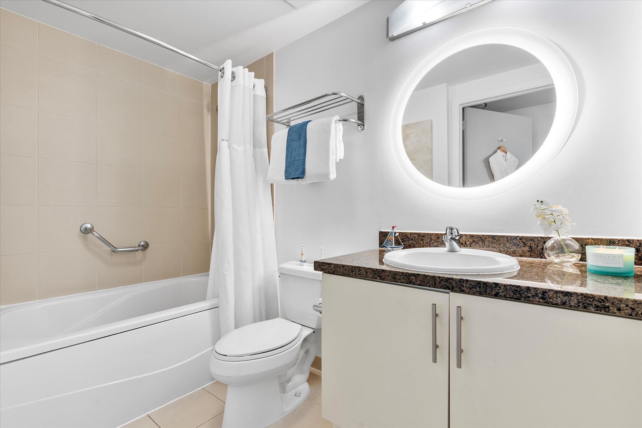 A full view of a clean, modern bathroom featuring a bathtub with a shower curtain, a toilet, and a vanity with a white sink and brown granite countertop. A large circular backlit mirror hangs above the sink.