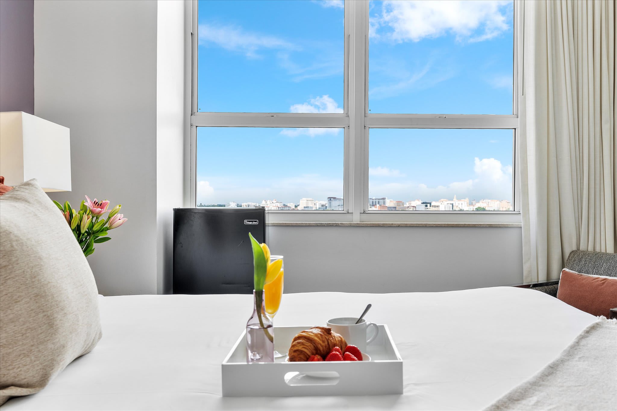 A close-up view of the white bed in a bright bedroom, showcasing a breakfast tray with a croissant, strawberries, and orange juice. A large window provides a city view in the background.