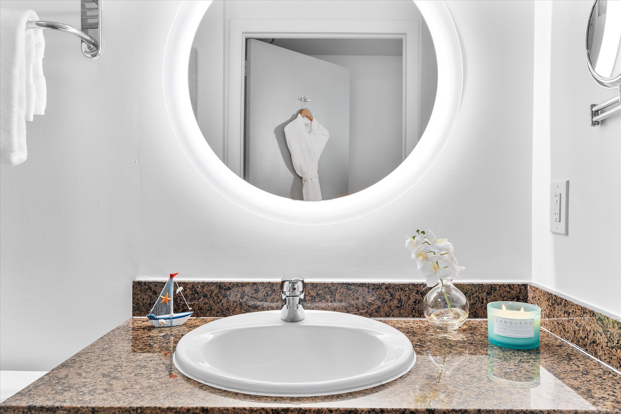 A modern bathroom vanity featuring a white oval sink set into a brown granite countertop, brightly illuminated by a large circular backlit mirror. A white bathrobe hangs on a hook, visible through the open door.