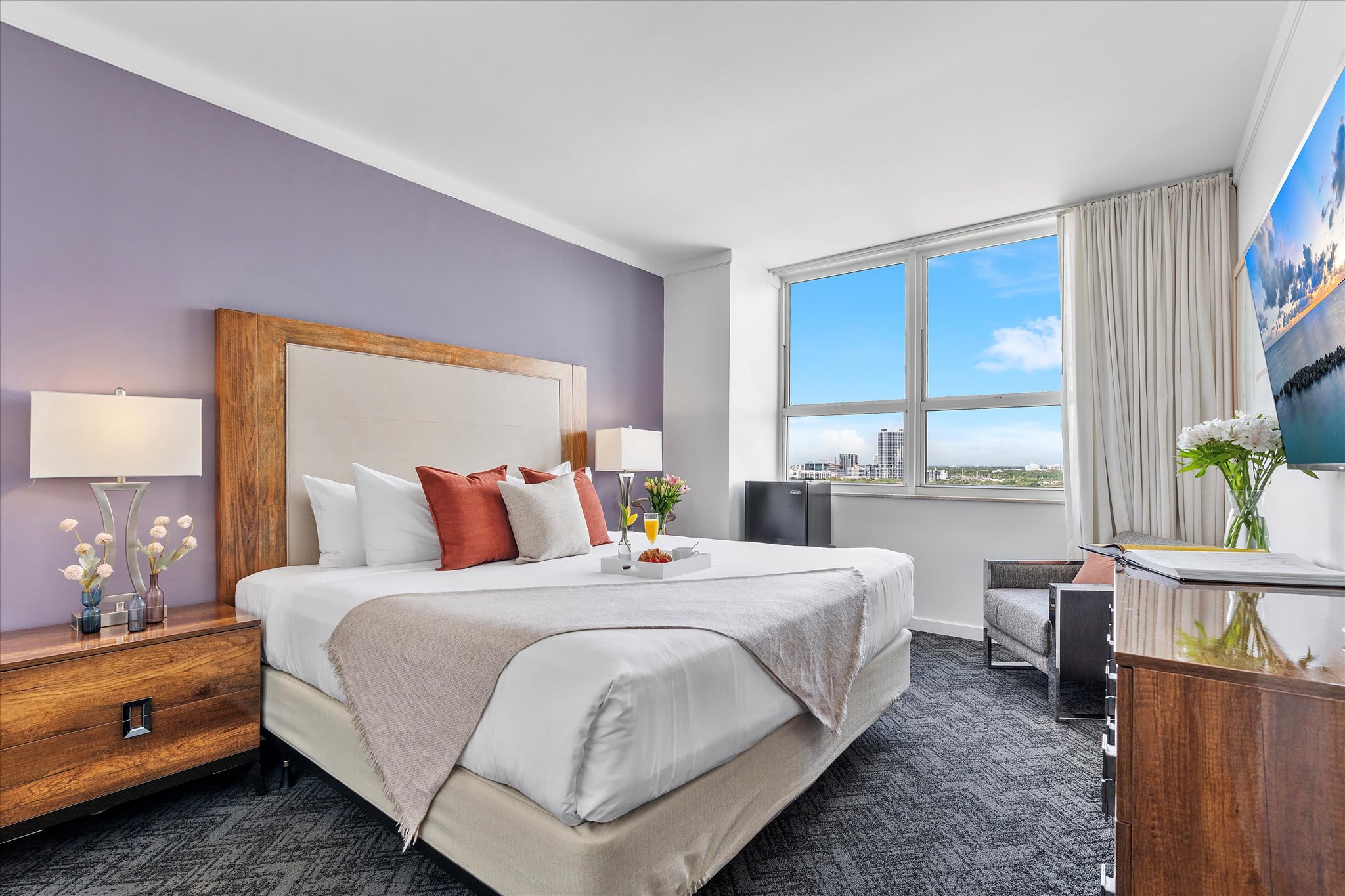 A modern bedroom featuring a king-sized bed with a large wooden and upholstered headboard, a purple accent wall, and dark patterned carpet. A large window offers a city view, and a flat-screen TV is mounted on the wall.